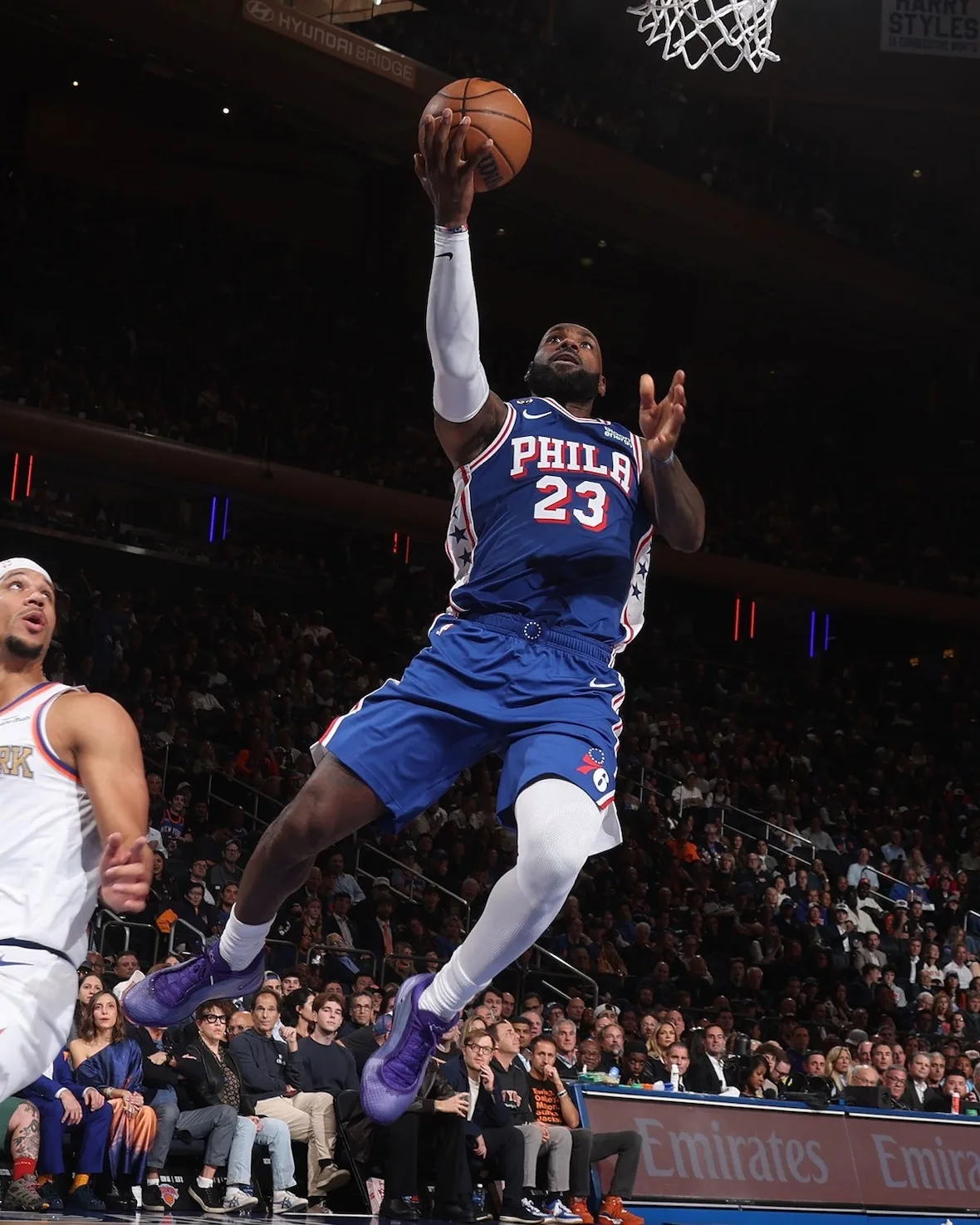 Player in blue Phila 23 jersey mid-air layup with purple sneakers, Emirates sign behind, Opening Night