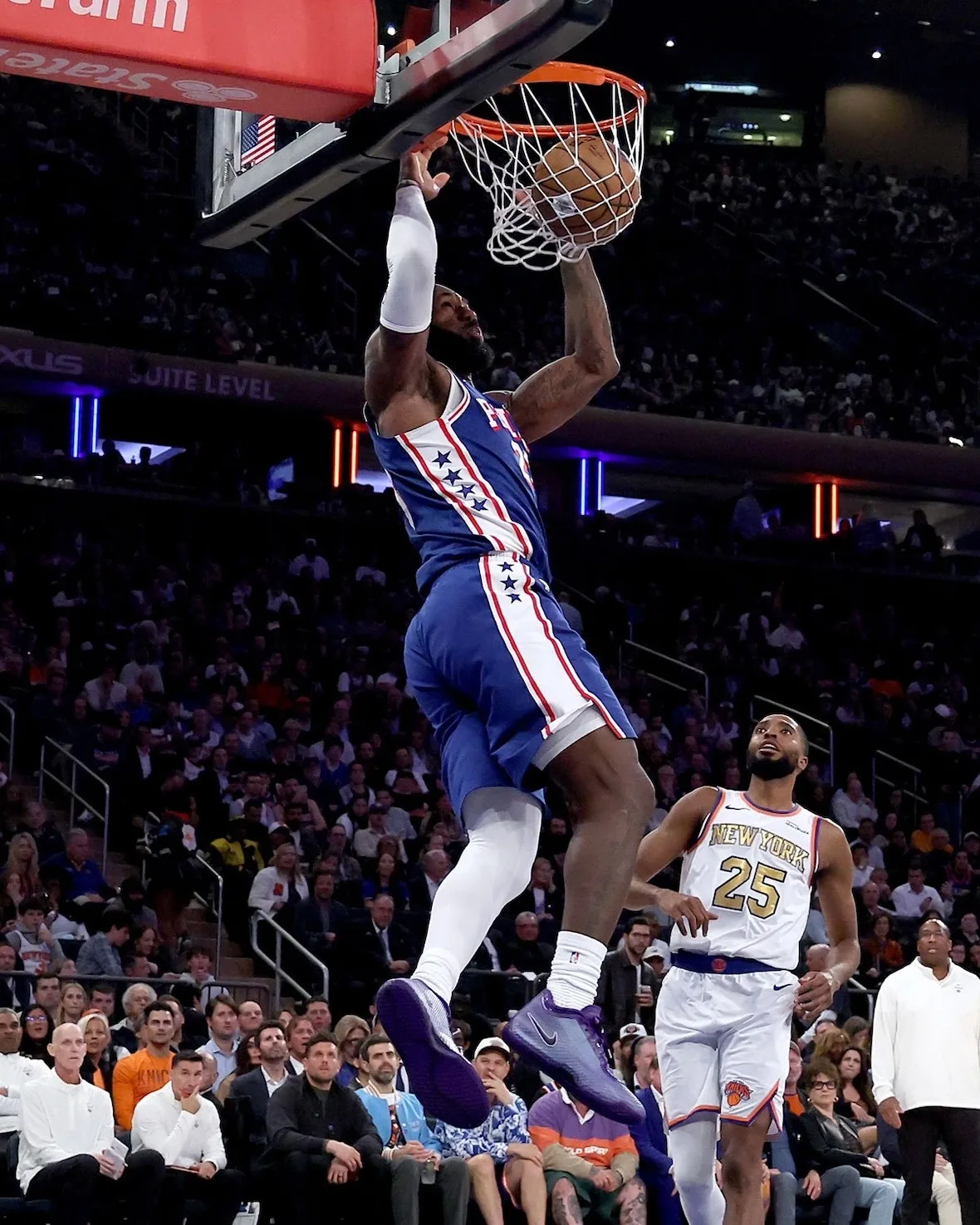 Player in blue Phila jersey dunking above rim, purple sneakers visible, arena crowd behind, Opening Night