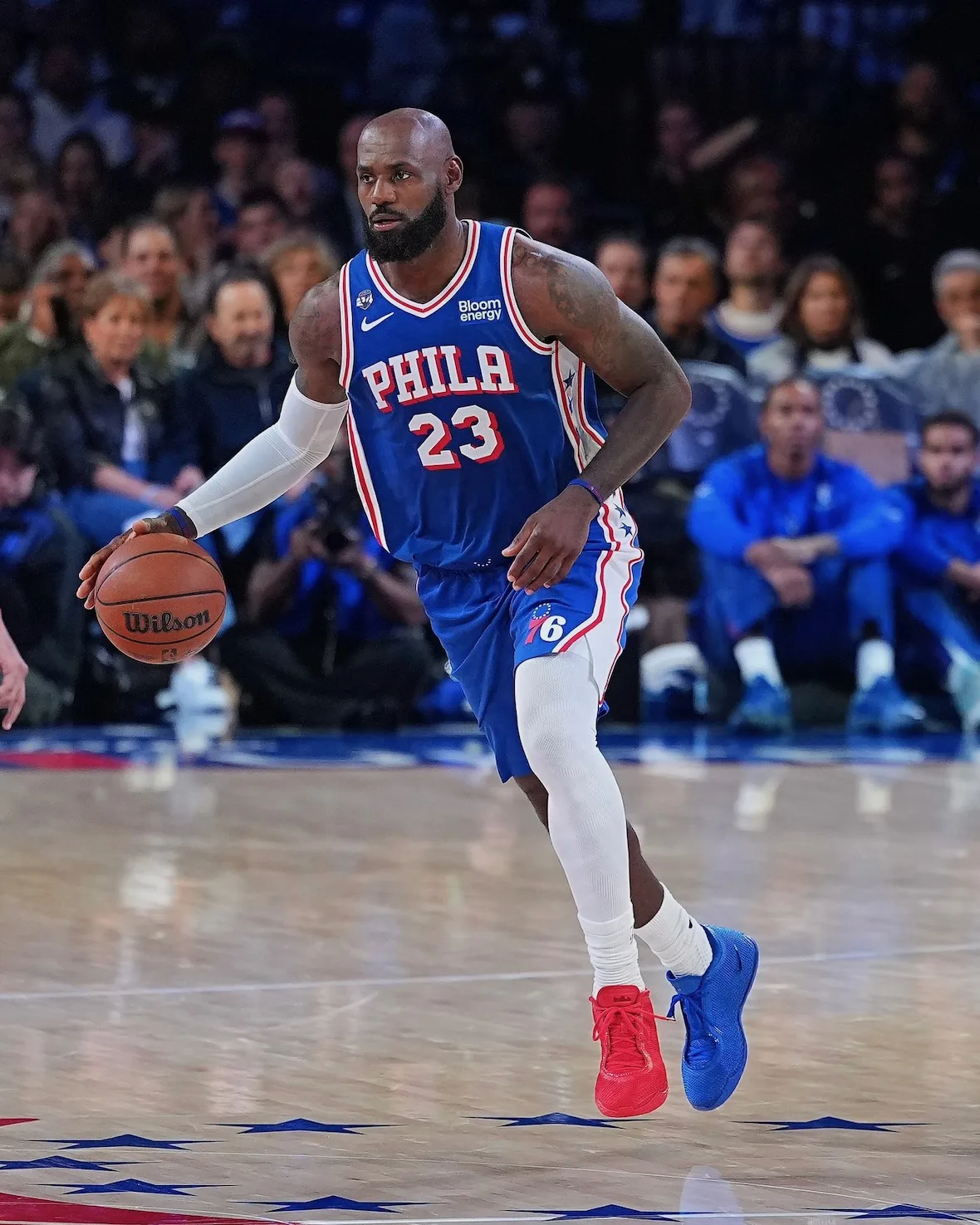 On-court dribble in blue Phila 23 jersey, red left shoe and blue right, star court logos, Philly