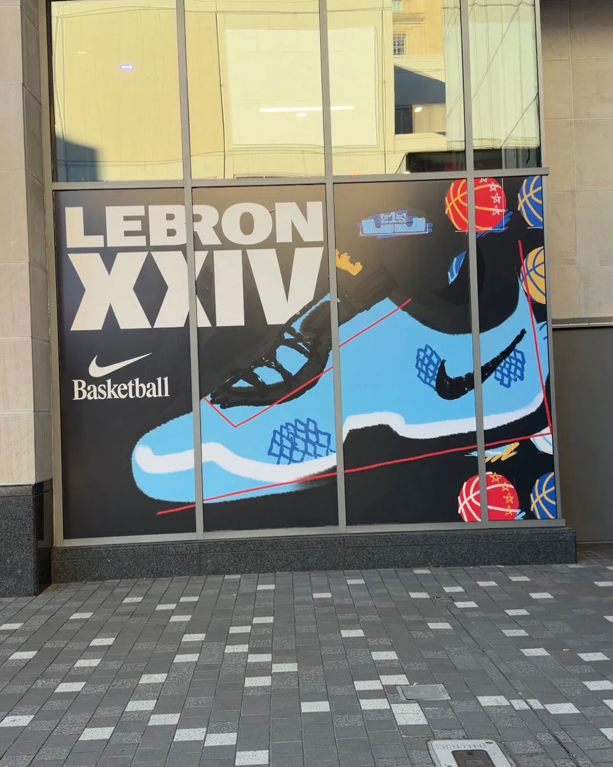 Storefront window ad reading LeBron XXIV and Nike Basketball, with light blue shoe artwork, Red and Blue