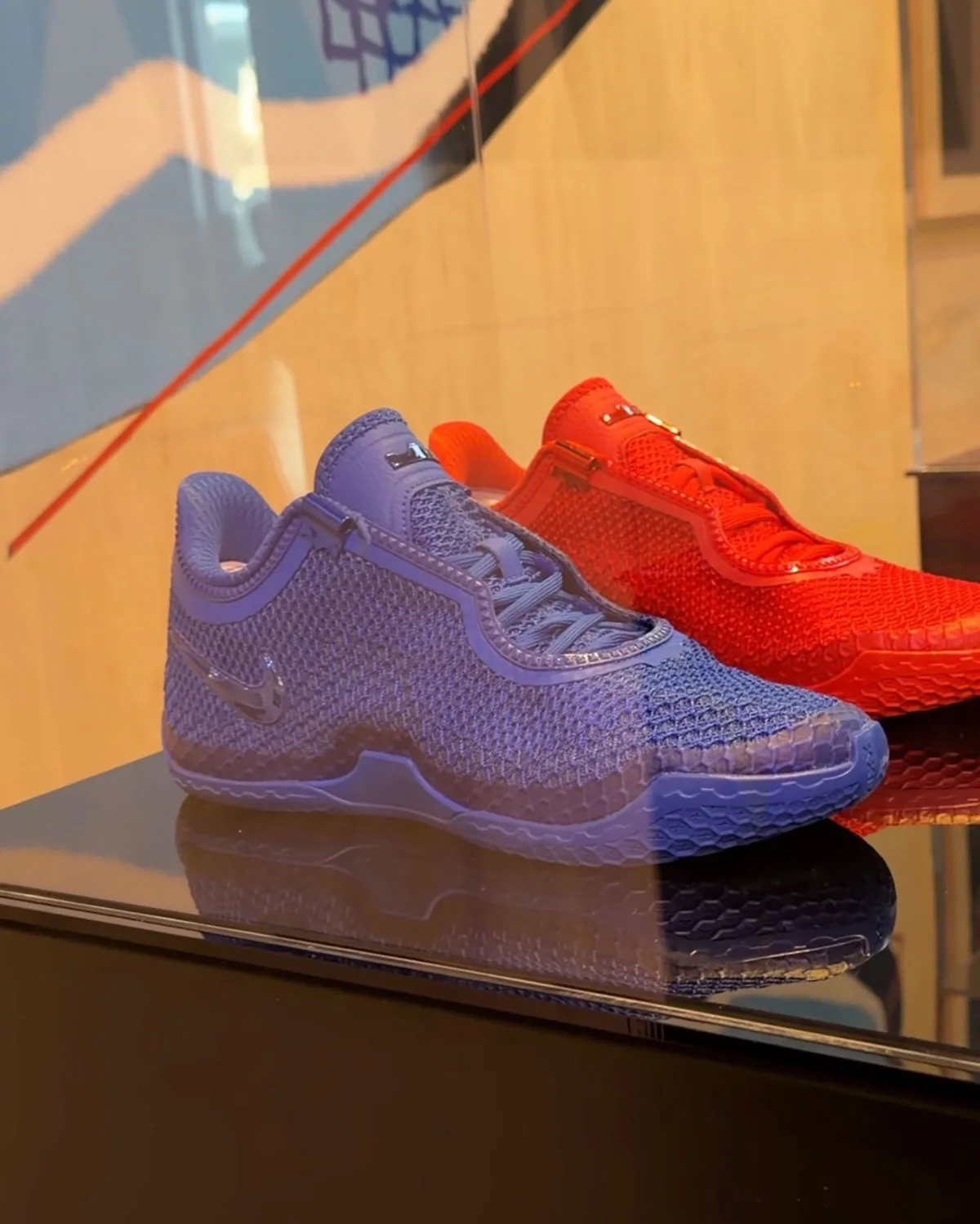 Lateral view of blue mesh shoe with glossy swoosh, red shoe behind, in display case, Red and Blue