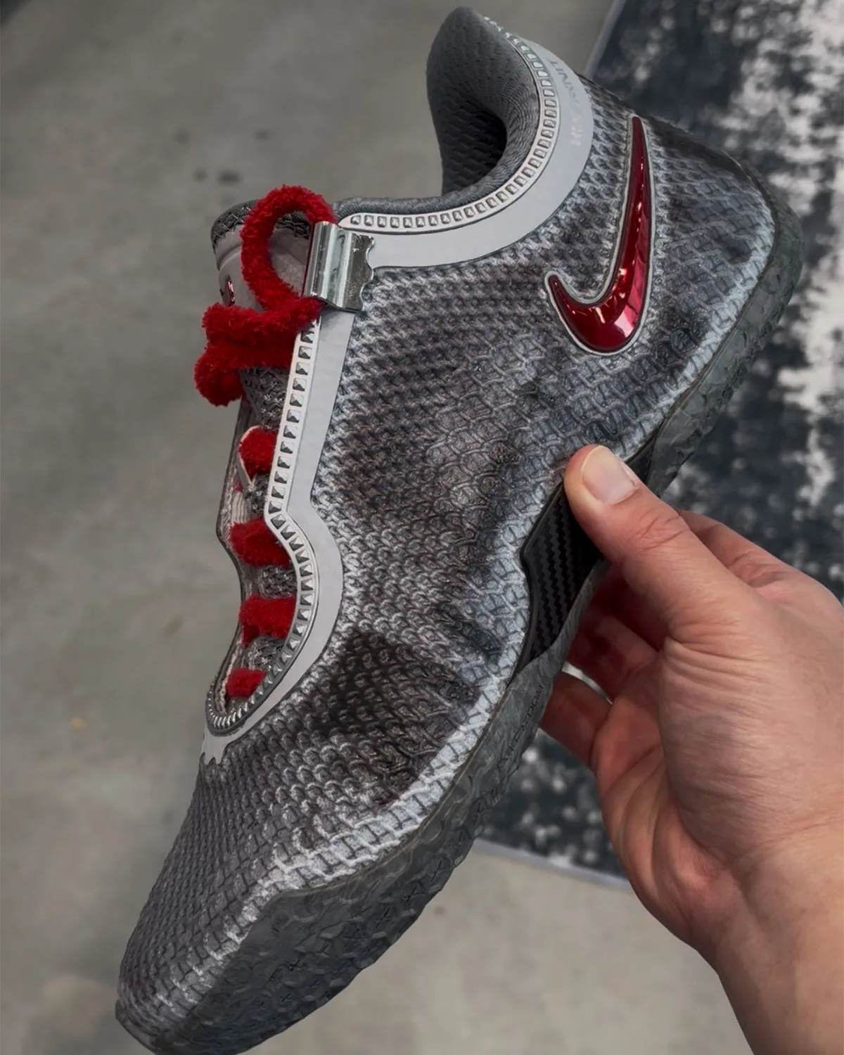Lateral view held in hand, grey scaled upper, red chrome swoosh, fuzzy red laces, concrete floor, Storm Drain