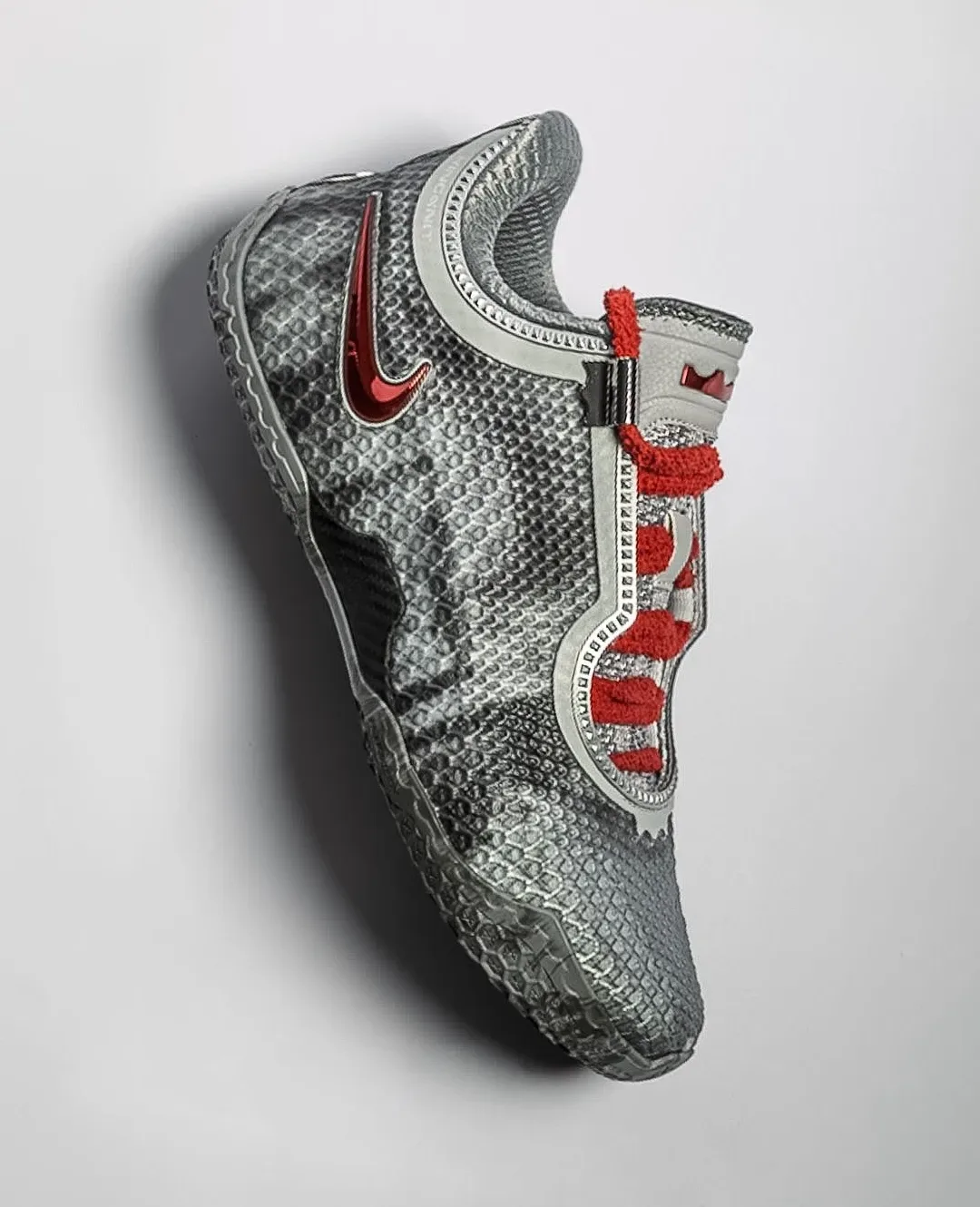 Lateral view on grey backdrop, snakeskin-textured grey upper, red chrome swoosh, fuzzy red laces, Storm Drain