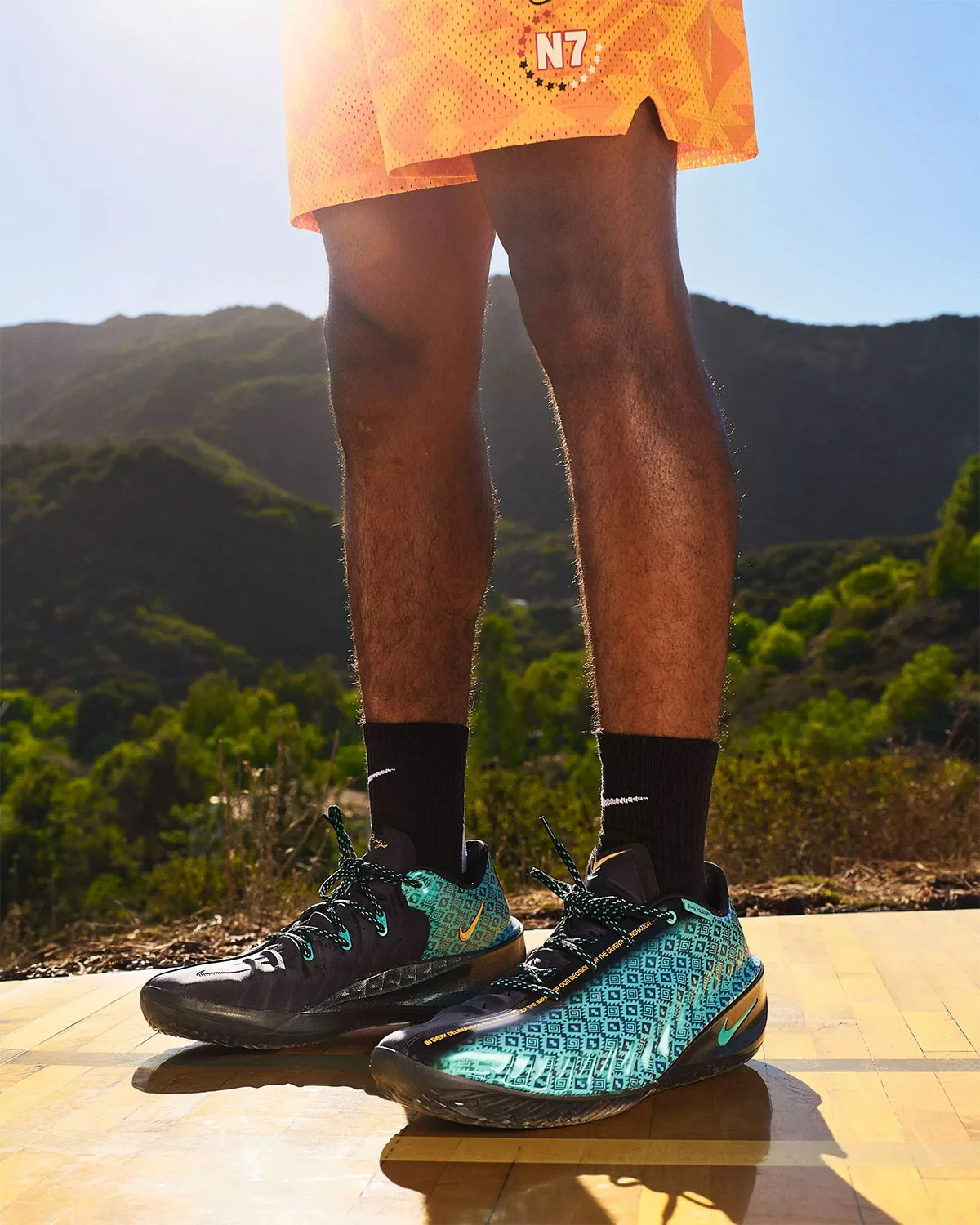 On-foot view, black and teal patterned basketball shoes, black socks, orange shorts, hills behind, Holiday 2026 Collection