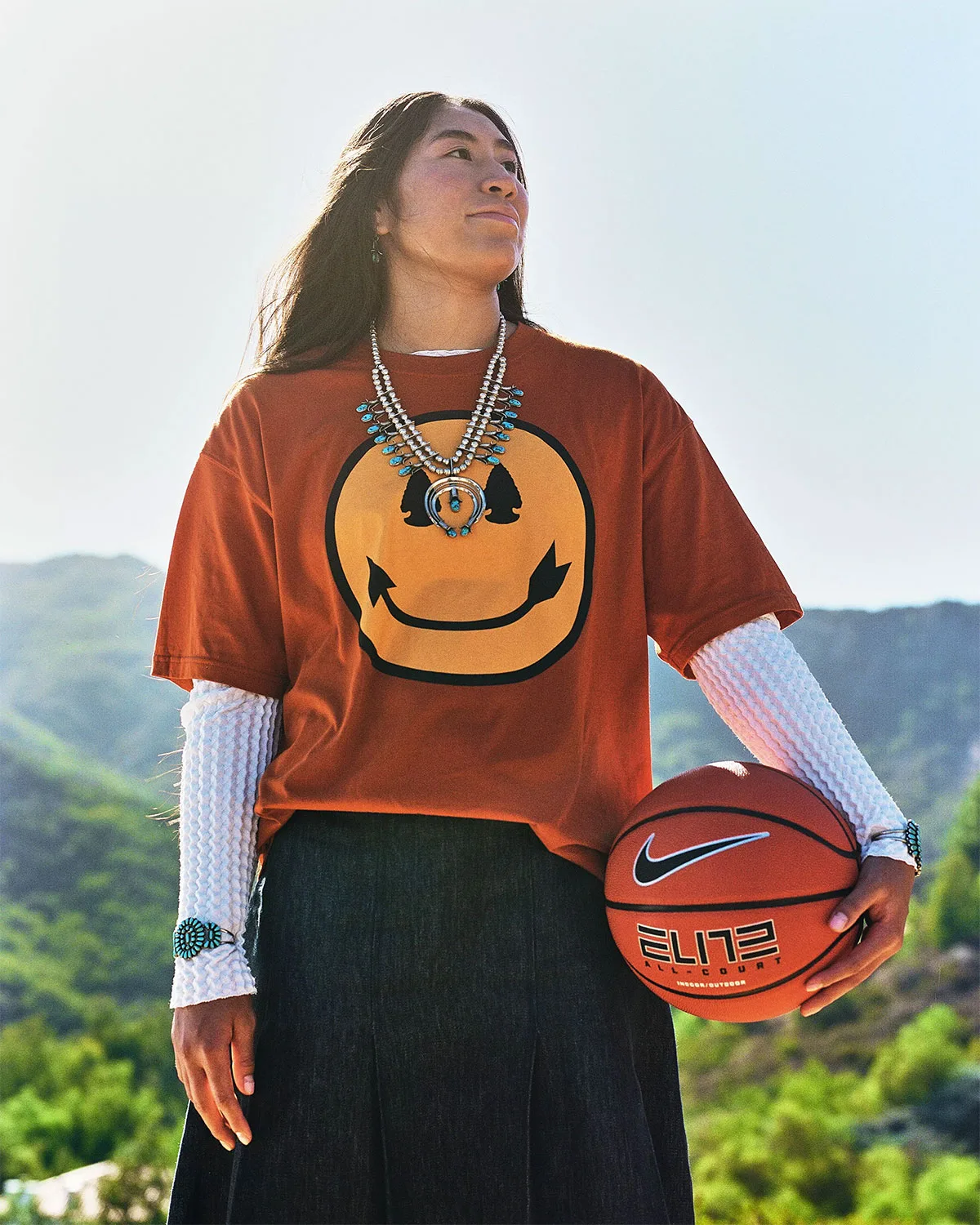 Woman holding Nike Elite basketball, orange tee with large smiley, silver and turquoise necklace, hills, Holiday 2026