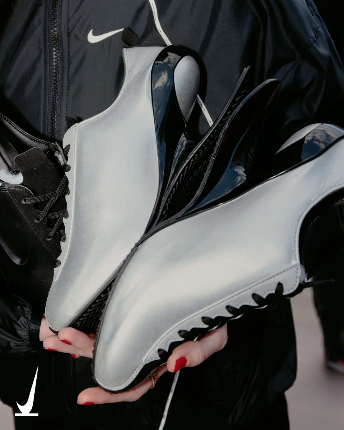 In-hand view of silver pair with black lacing and glossy black curved heels, dark jacket behind, Nike Ovolo 2.0