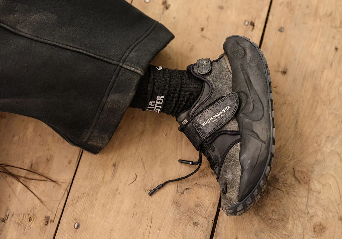 On-foot view with a branded dark strap, black trousers and wooden flooring, Nike Rift Rocker