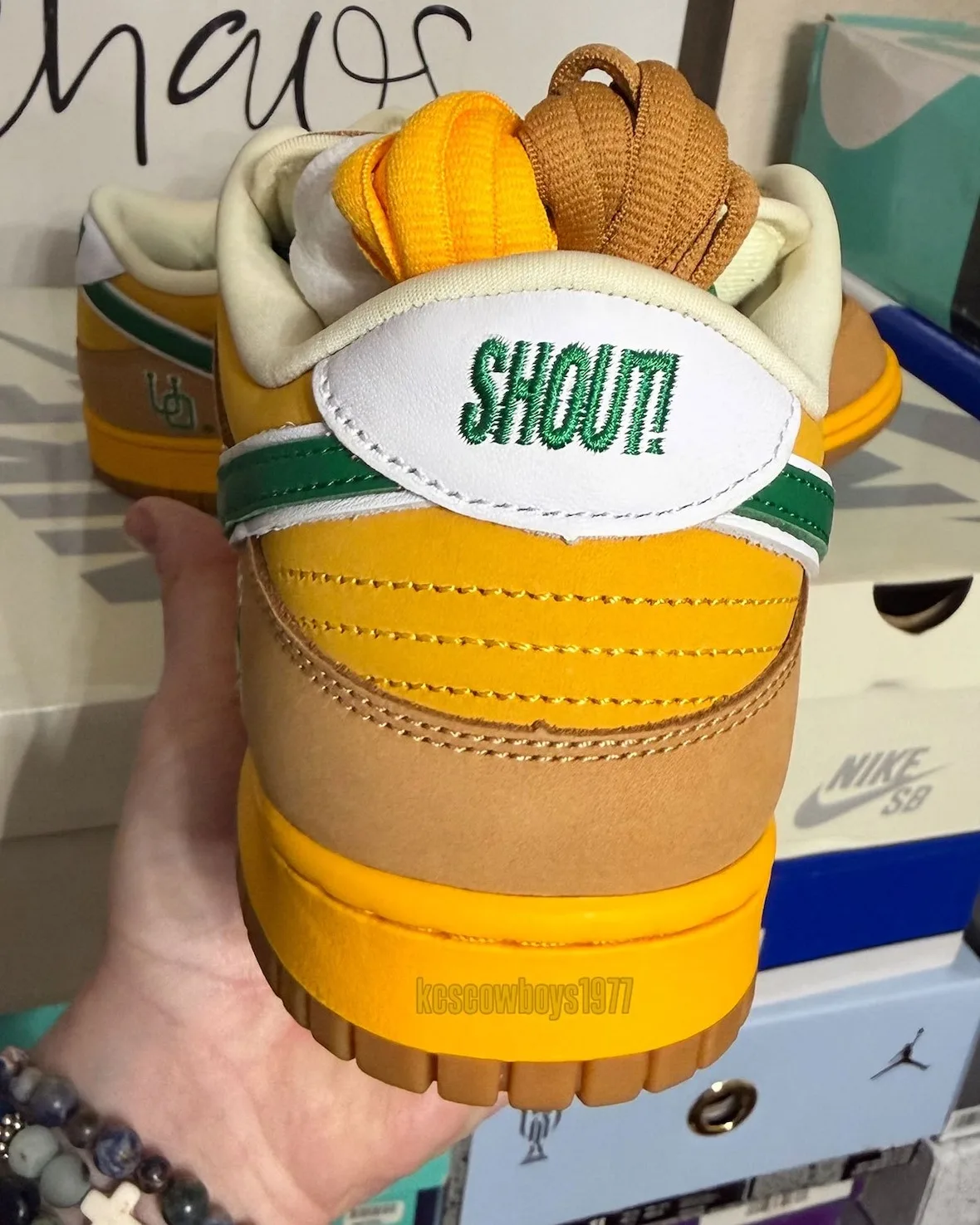 Heel view in hand, white tab with green SHOUT! embroidery, yellow and tan laces, Shout