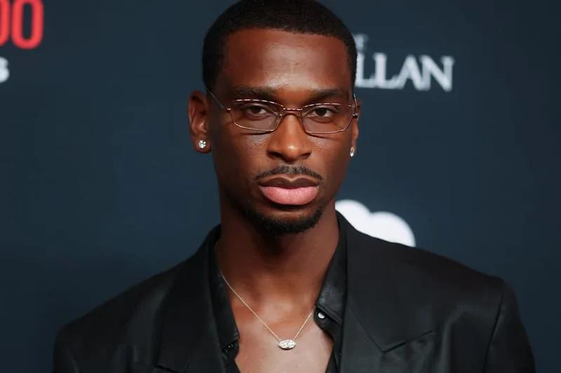Portrait of a man in glasses, black jacket and pendant necklace in front of a dark step-and-repeat backdrop, Nike SHAI 002