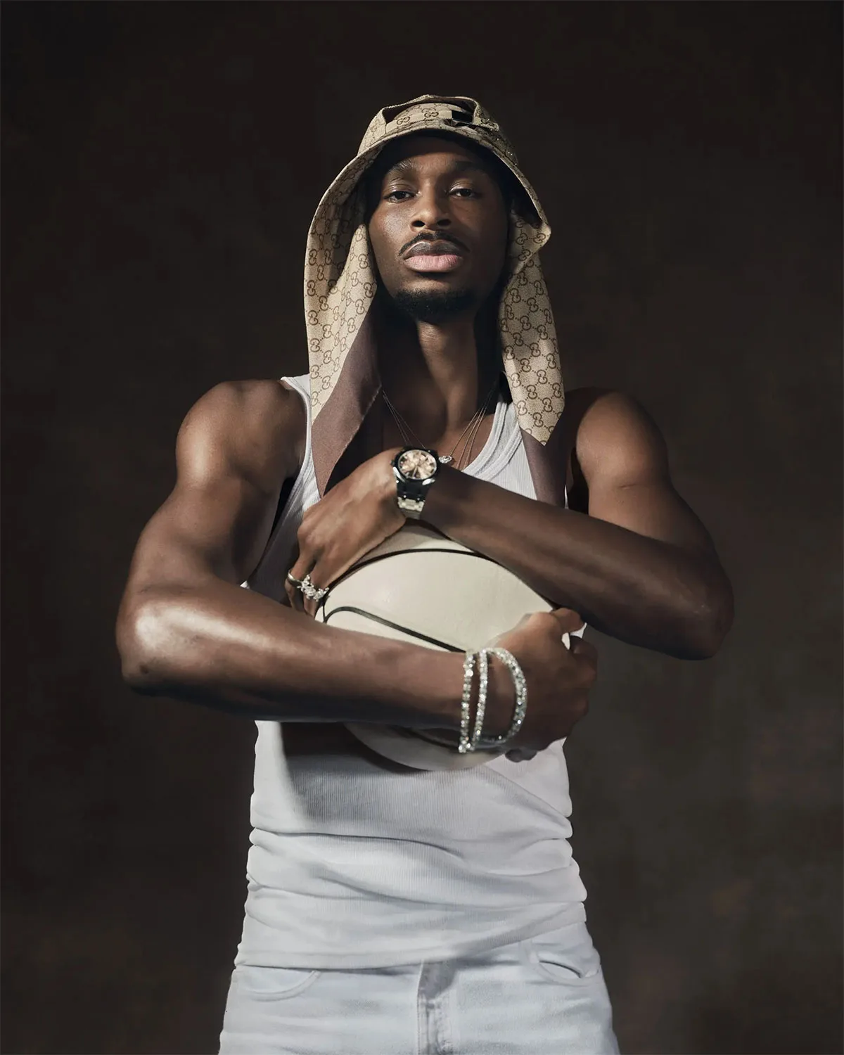 Full-length studio portrait: man in monogram bucket hat, white tank and jeans hugging a cream basketball, jewelled