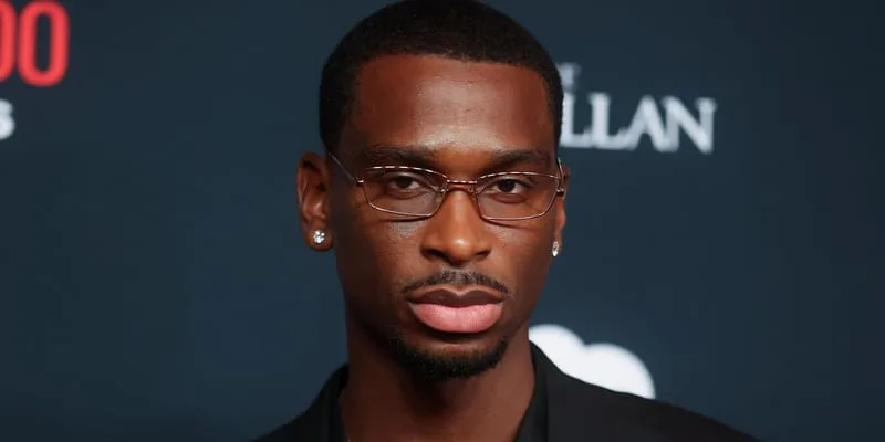 Close-up portrait of a man in thin-rimmed glasses and stud earrings against a dark event backdrop with partial text, Nike