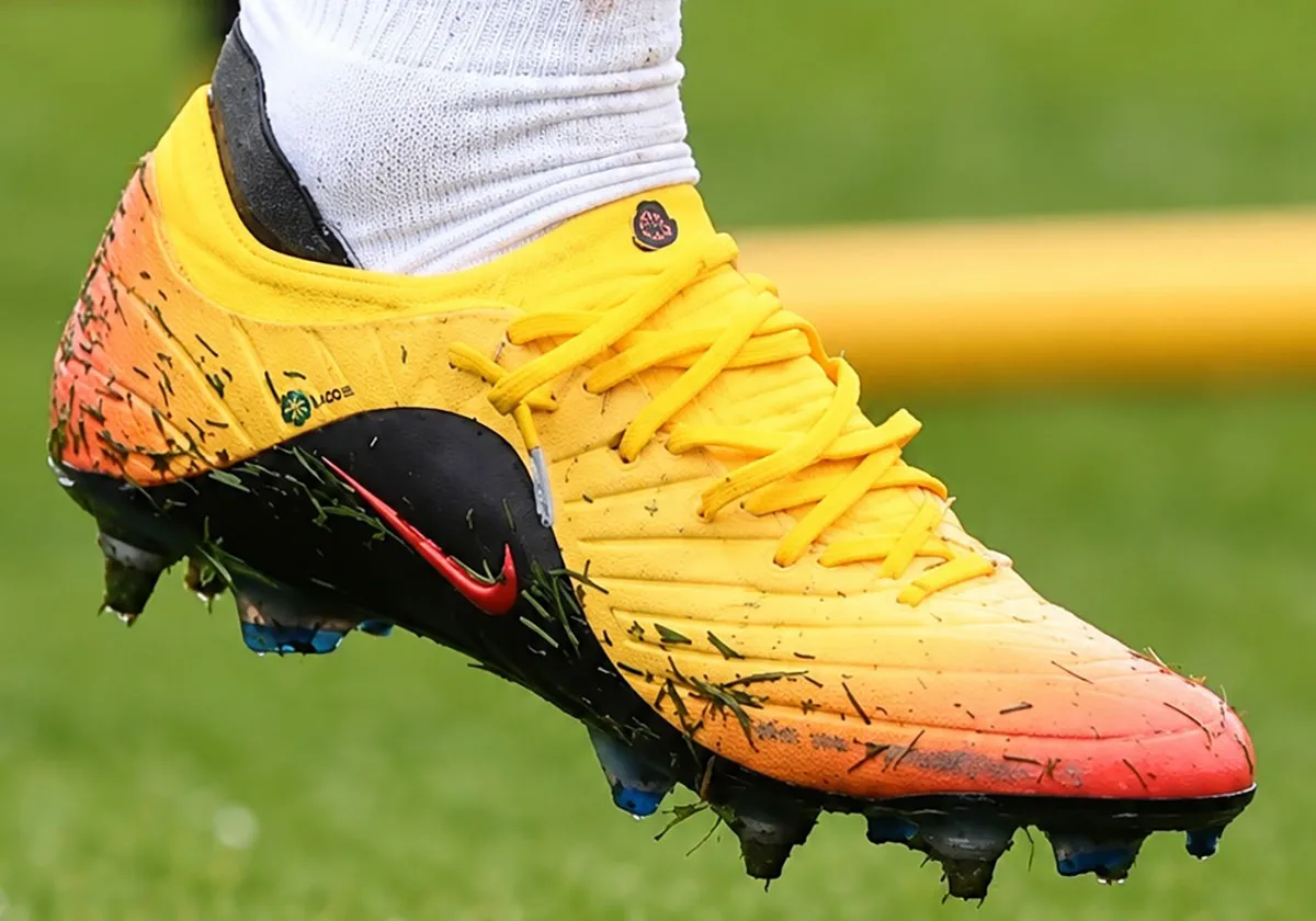 On-foot lateral view of yellow-to-orange boot with grass specks, red swoosh and yellow laces, Sundial