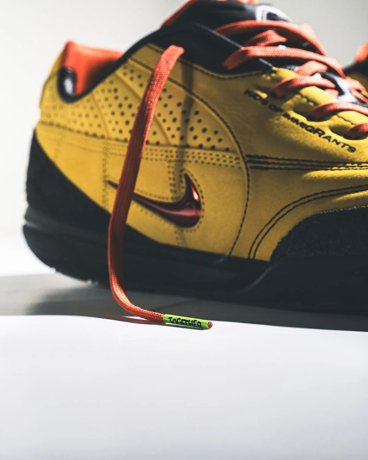 Low-angle lateral detail of yellow perforated suede, red chrome swoosh and orange lace with green "Together" tip, Amarillo