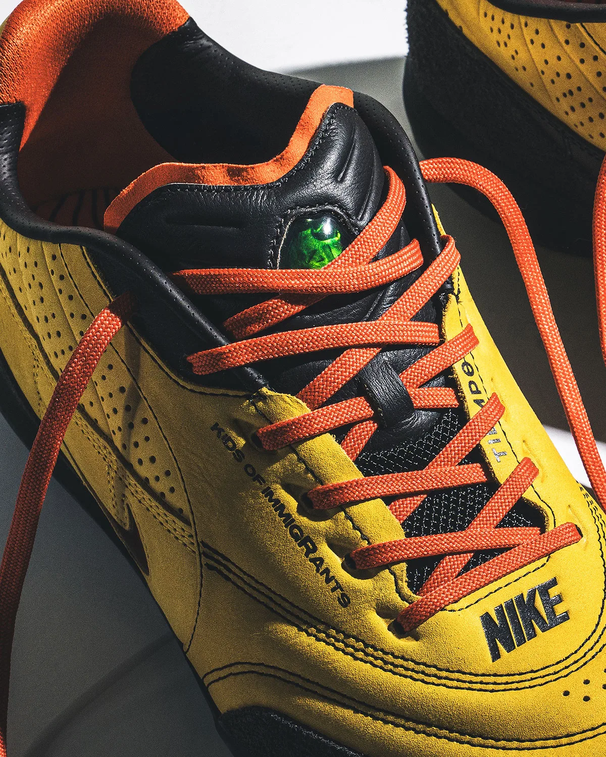 Close-up top-down of yellow suede, orange laces, black leather tongue with green gem and Nike text, Amarillo