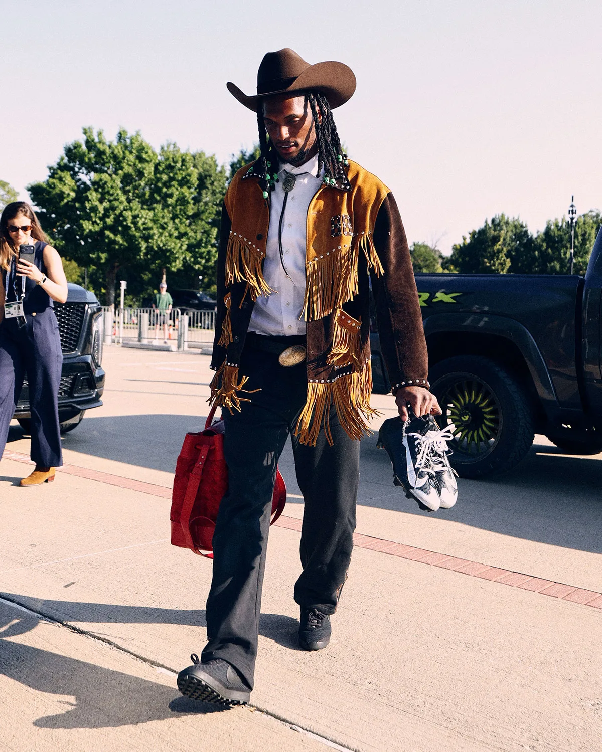 On-foot shot of man in fringed suede jacket carrying red bag and black and white cleats in hand, Vapor Pro 1