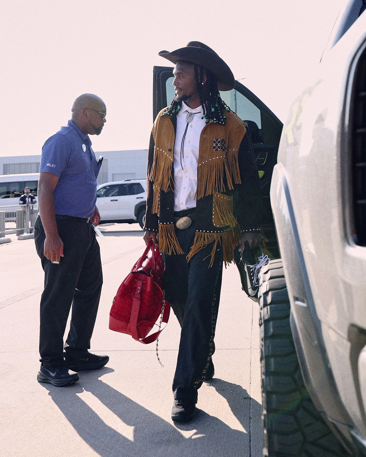 On-foot shot of man in cowboy hat and fringed jacket holding black cleats with a white swoosh by a truck, Vapor Pro 1