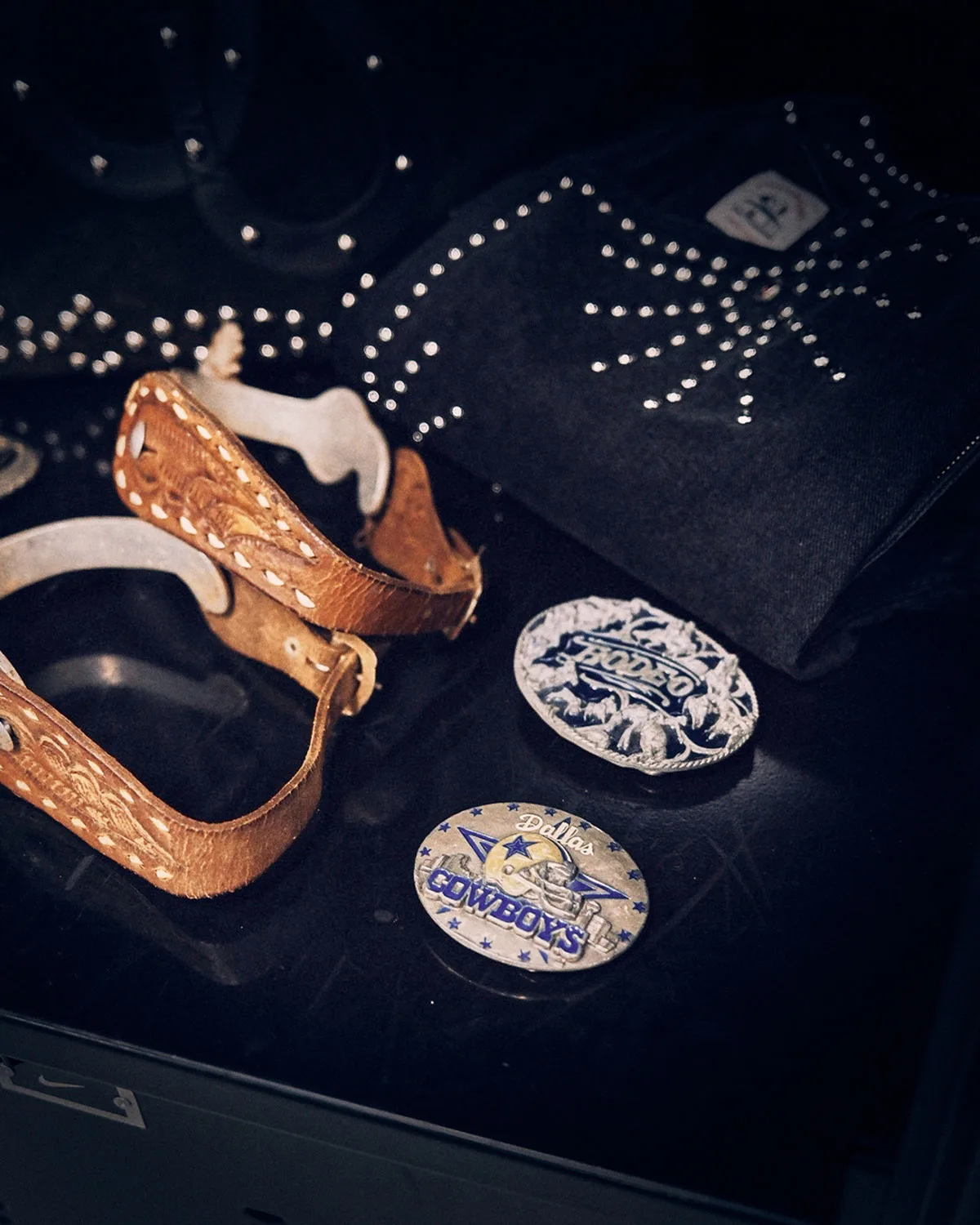 Detail of leather spurs and Rodeo and Dallas Cowboys belt buckles beside studded black fabric, Vapor Pro 1