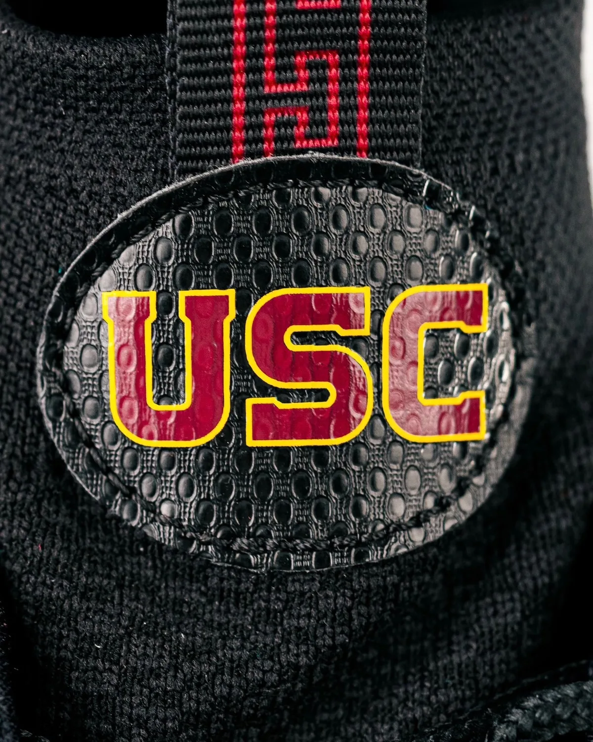 Close-up of oval USC patch in maroon and gold on black textured upper, with red patterned strap, Pro USC PE