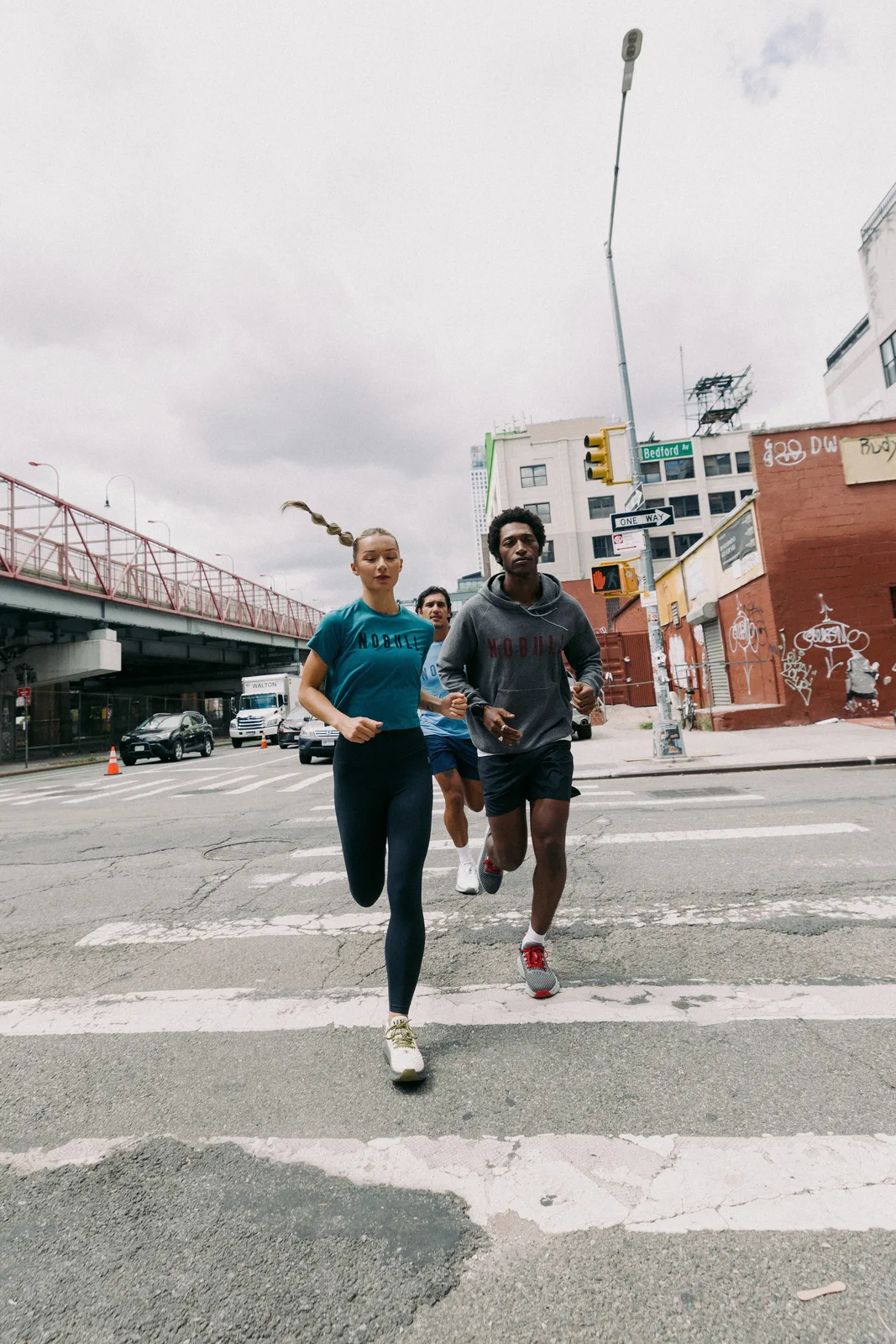 Three runners crossing a city street, shoes in grey and red, cream, and white, NOBULL Journey 2