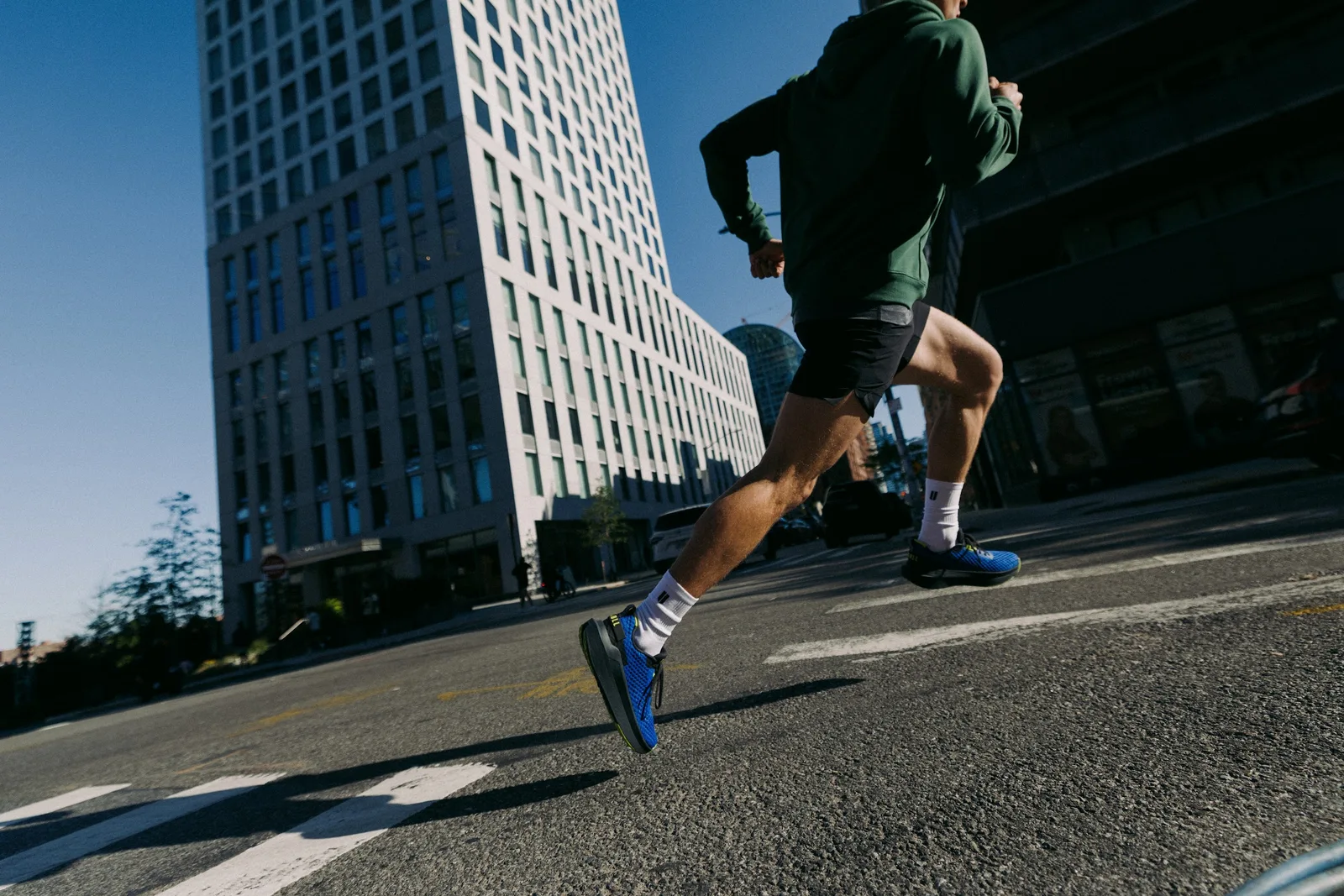 Low-angle runner in green hoodie mid-stride, royal blue shoes with lime trim, tall building behind, NOBULL Journey 2