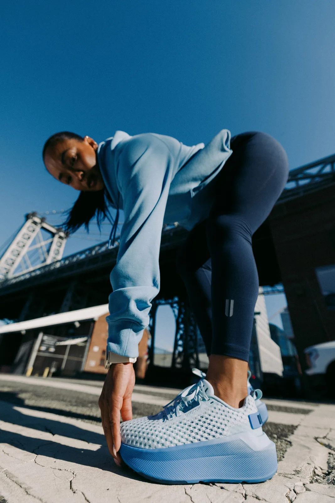 Low-angle on-foot, person stretching in light blue mesh shoes with blue midsole, bridge and sky behind, NOBULL Journey 2