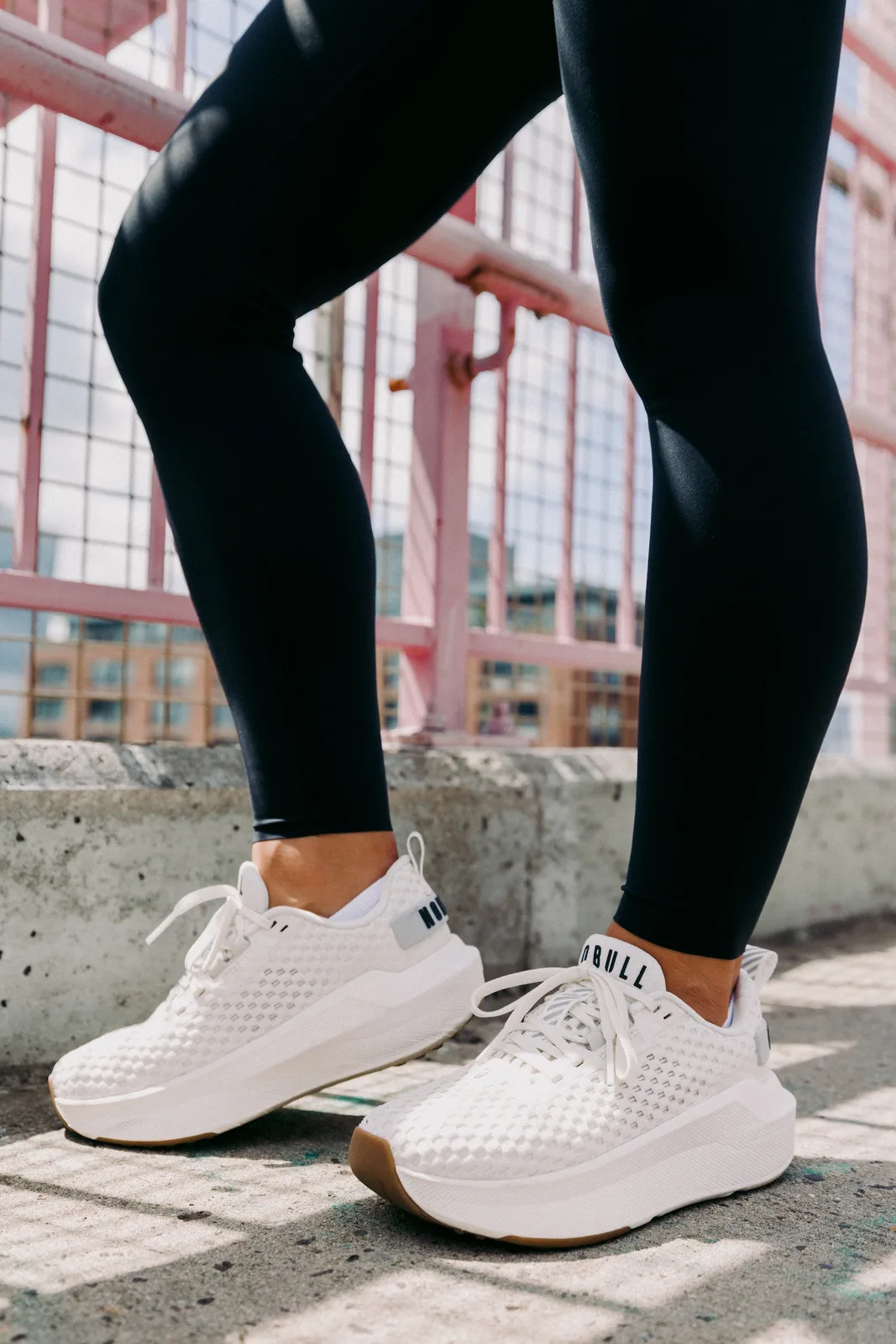 Lateral on-foot view of white mesh shoes with gum soles, black leggings, pink railing behind, NOBULL Journey 2
