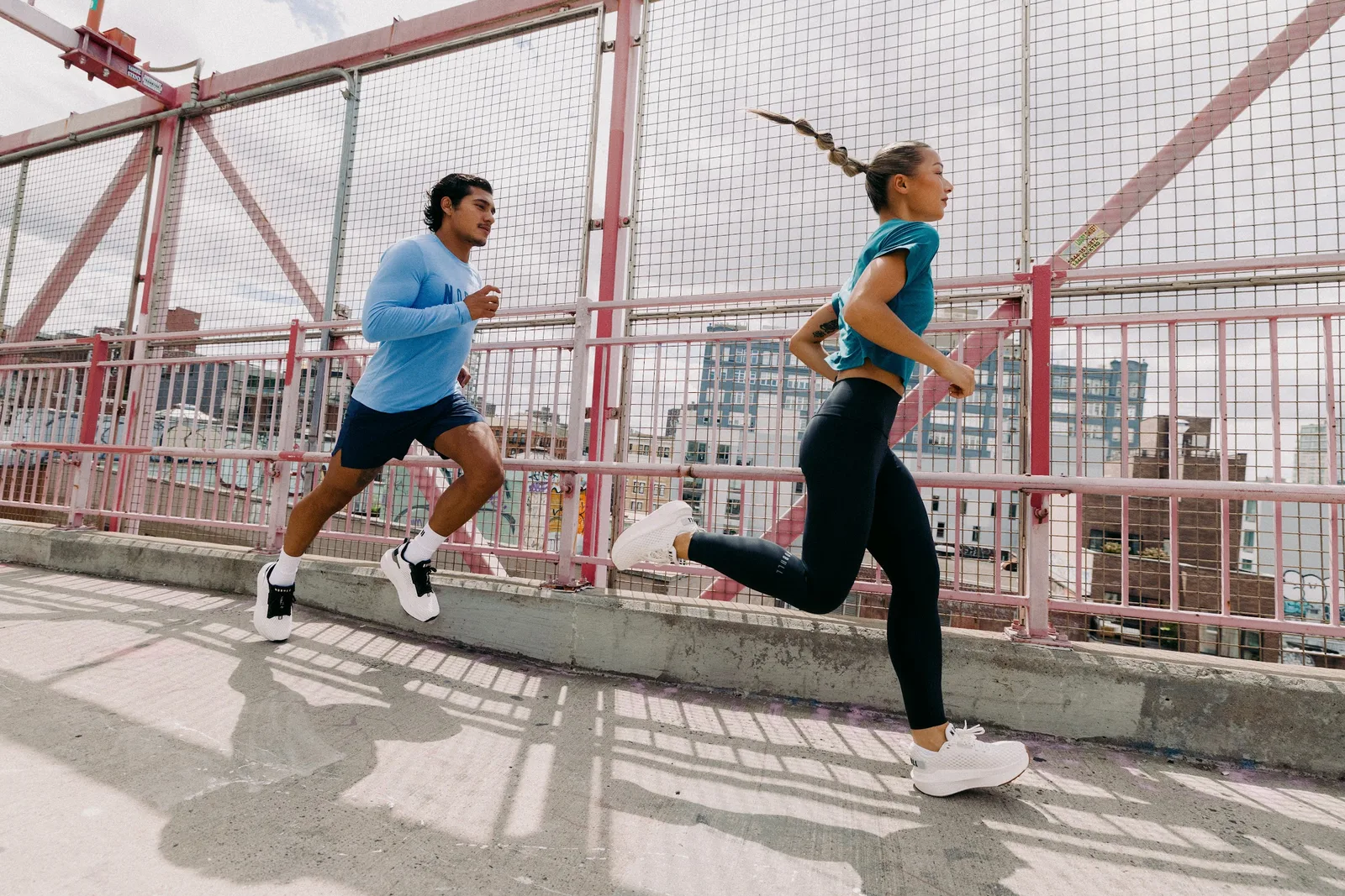 Two runners mid-stride on a pink-railed bridge, both in white mesh running shoes, NOBULL Journey 2