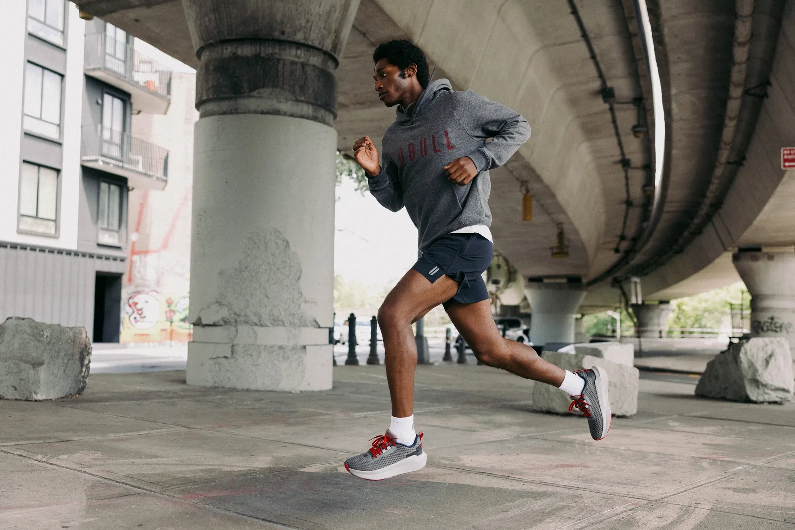 Runner in grey NOBULL hoodie mid-stride under an overpass, grey shoes with red laces, NOBULL Journey 2