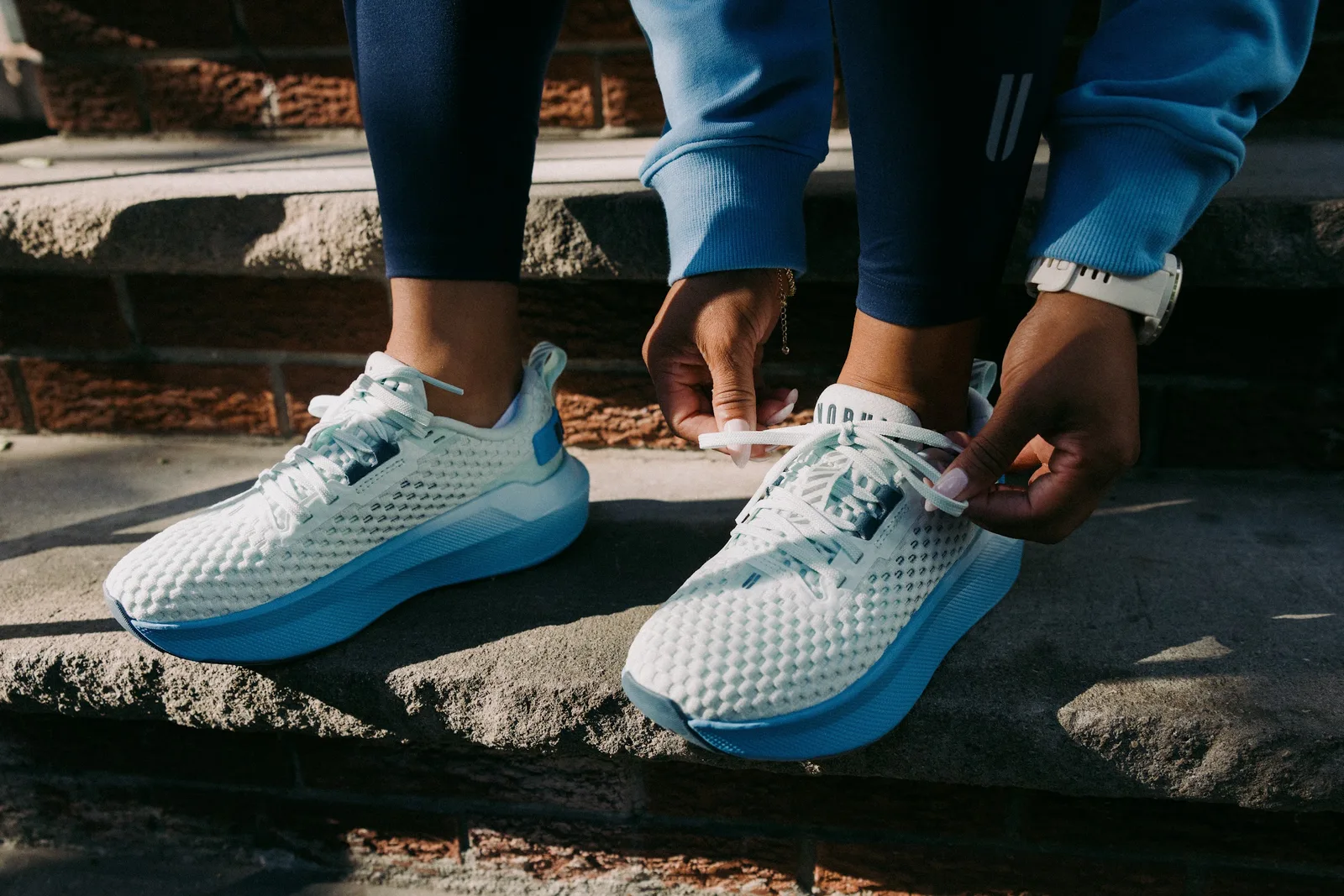 On-foot, hands tying laces on white mesh shoes with blue midsoles, brick steps behind, NOBULL Journey 2