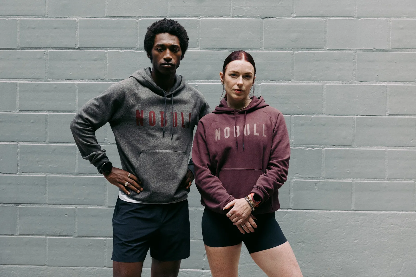 Two people in NOBULL hoodies, grey with red logo and burgundy, against a pale green brick wall, NOBULL Journey 2