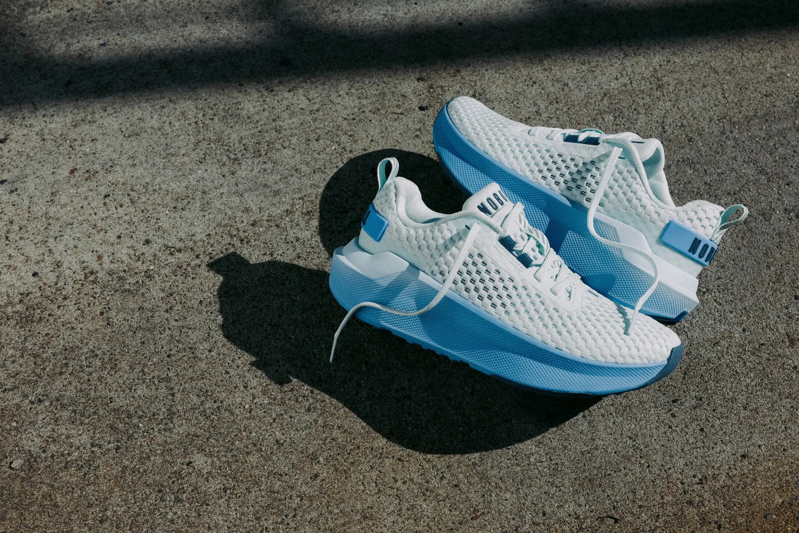 Pair on rough concrete, white mesh uppers with light blue midsoles and blue heel tab with NOBULL text, NOBULL Journey 2