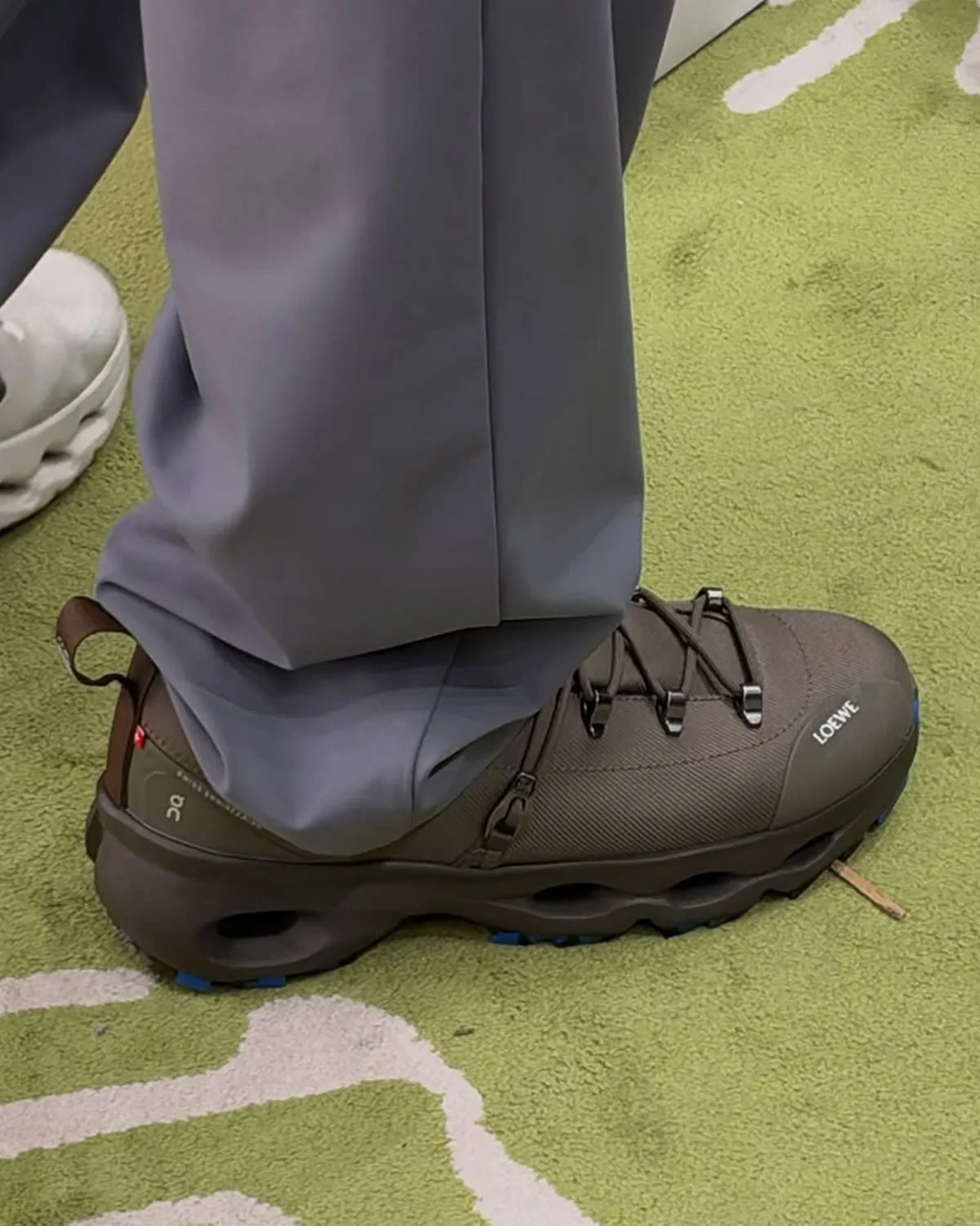 On-foot lateral view, brown shoe with heel pull loop, cutout midsole, blue outsole accents, green rug, Hiker x Loewe