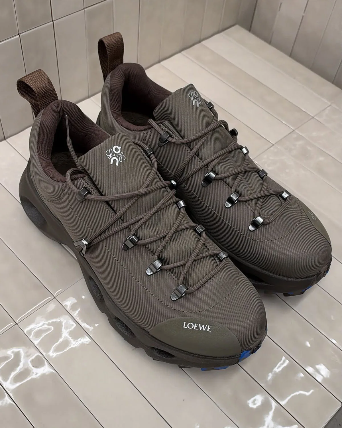 Top-down pair on tiled floor, brown ribbed fabric, metal lace hooks, white LOEWE toe text, Hiker x Loewe