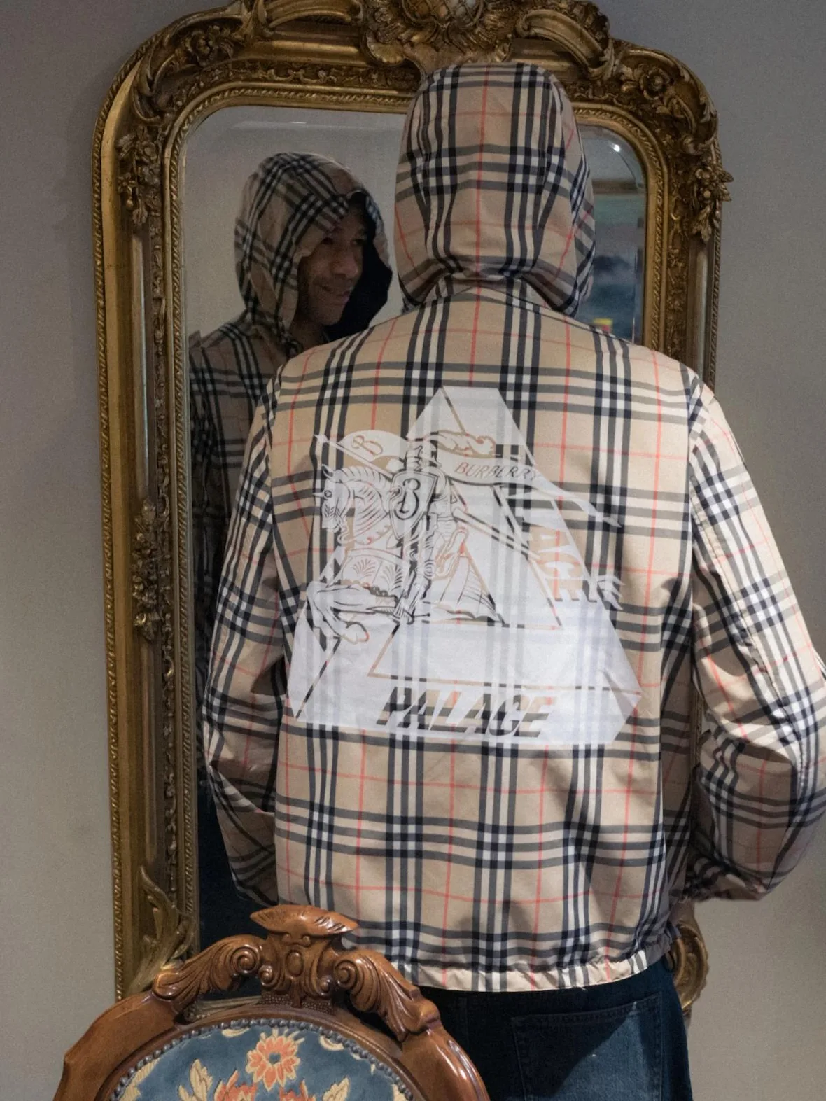 Rear on-body view of hooded beige check jacket with white Palace triangle graphic, mirror and carved chair, x Burberry