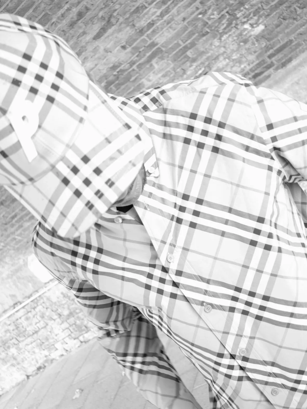 Black and white on-body detail of oversized check shirt and check cap against a brick wall, x Burberry Collection