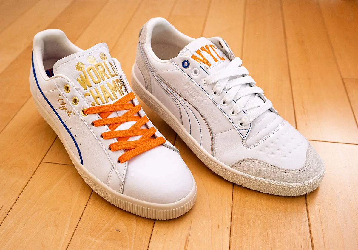 Pair top-down: white sneaker with gold "World Champs" tongue and orange laces beside one with orange NYC tongue, Ceremony