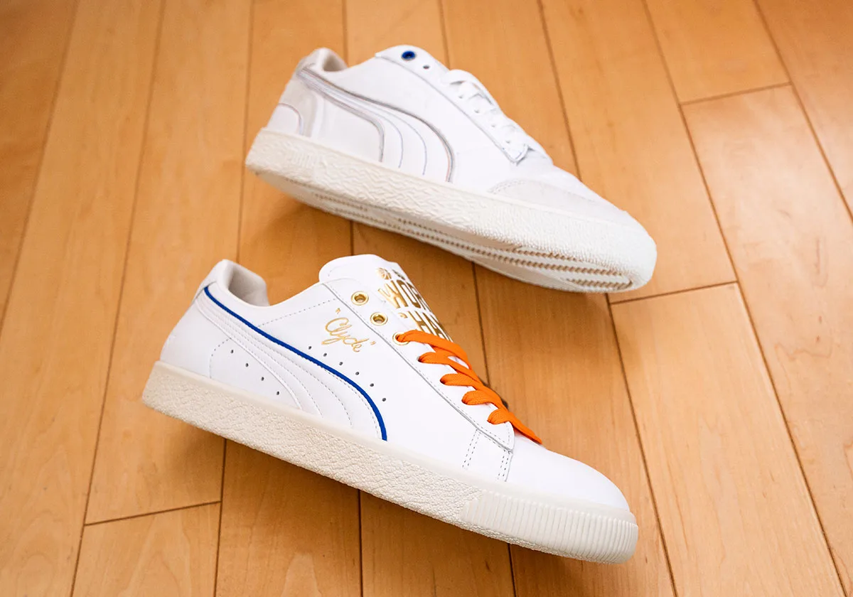 Pair on wood floor: white sneaker with orange laces and blue stripe beside a white suede-trimmed pair, Ceremony
