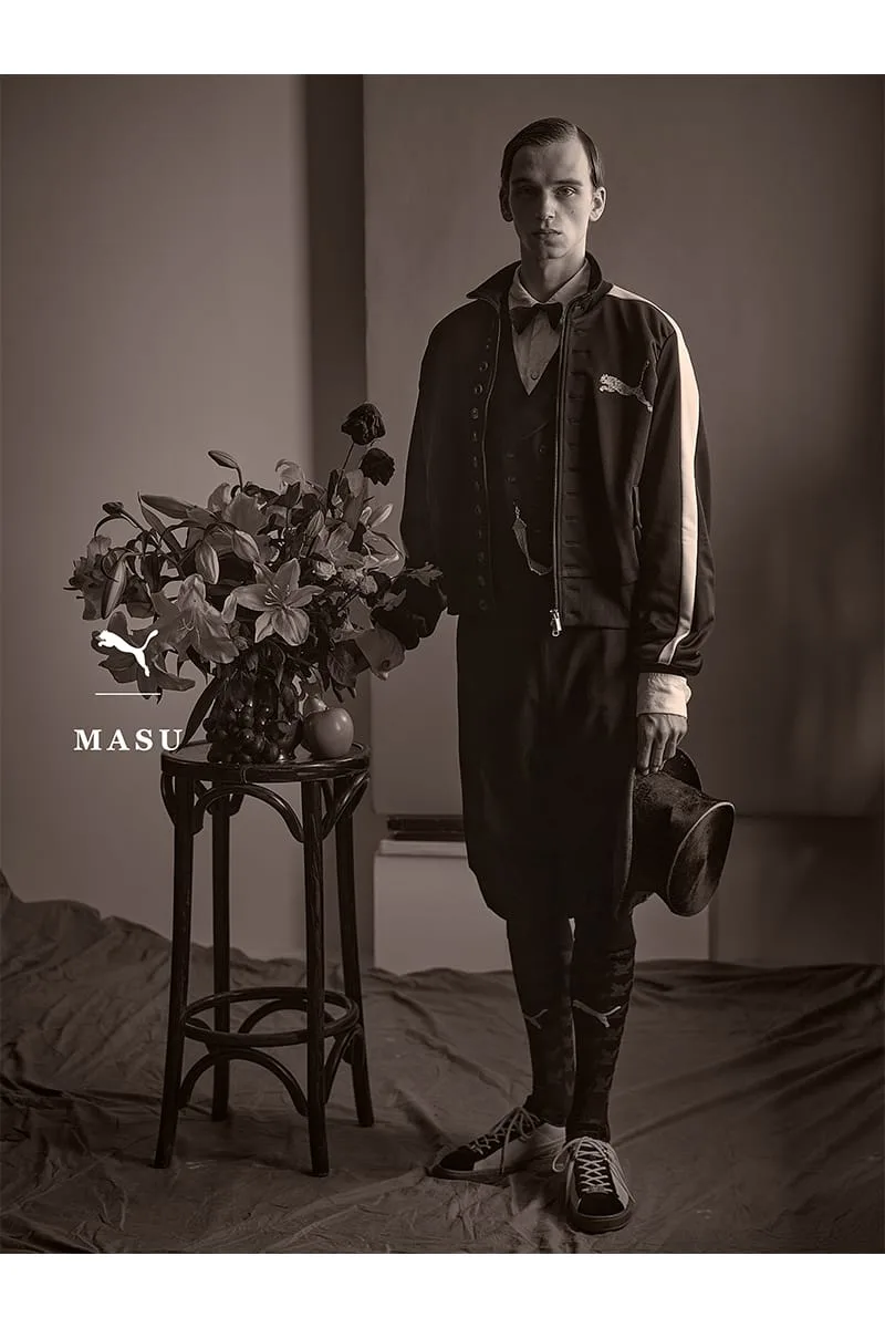 Sepia full-length shot of model in jacket and bow tie beside flowers, holding top hat, IVORYxBLACK