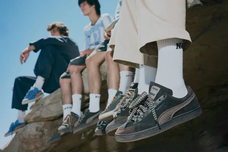 Low-angle on-foot view of people sitting on a rock ledge, dark suede pair with patterned laces in front, NAHMIAS Suede