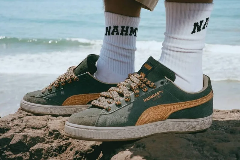 On-foot lateral view of green suede pair with orange crochet stripe, white NAHMIAS socks, ocean behind, NAHMIAS Suede Crochet