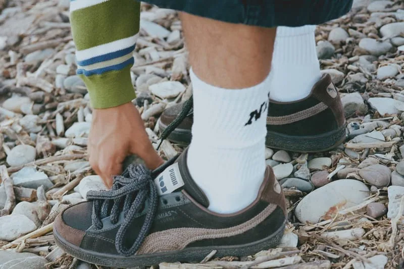 On-foot view of dark brown pair with grey laces, a hand adjusting the heel on a pebbly beach, NAHMIAS Suede Crochet
