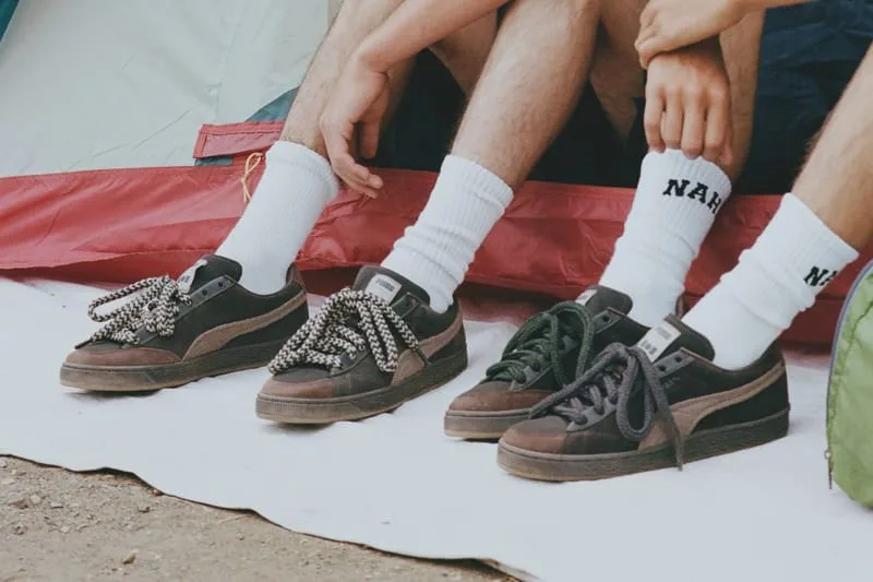 On-foot row of dark brown pairs with white socks printed with black text, seated by a red tent, NAHMIAS Suede Crochet