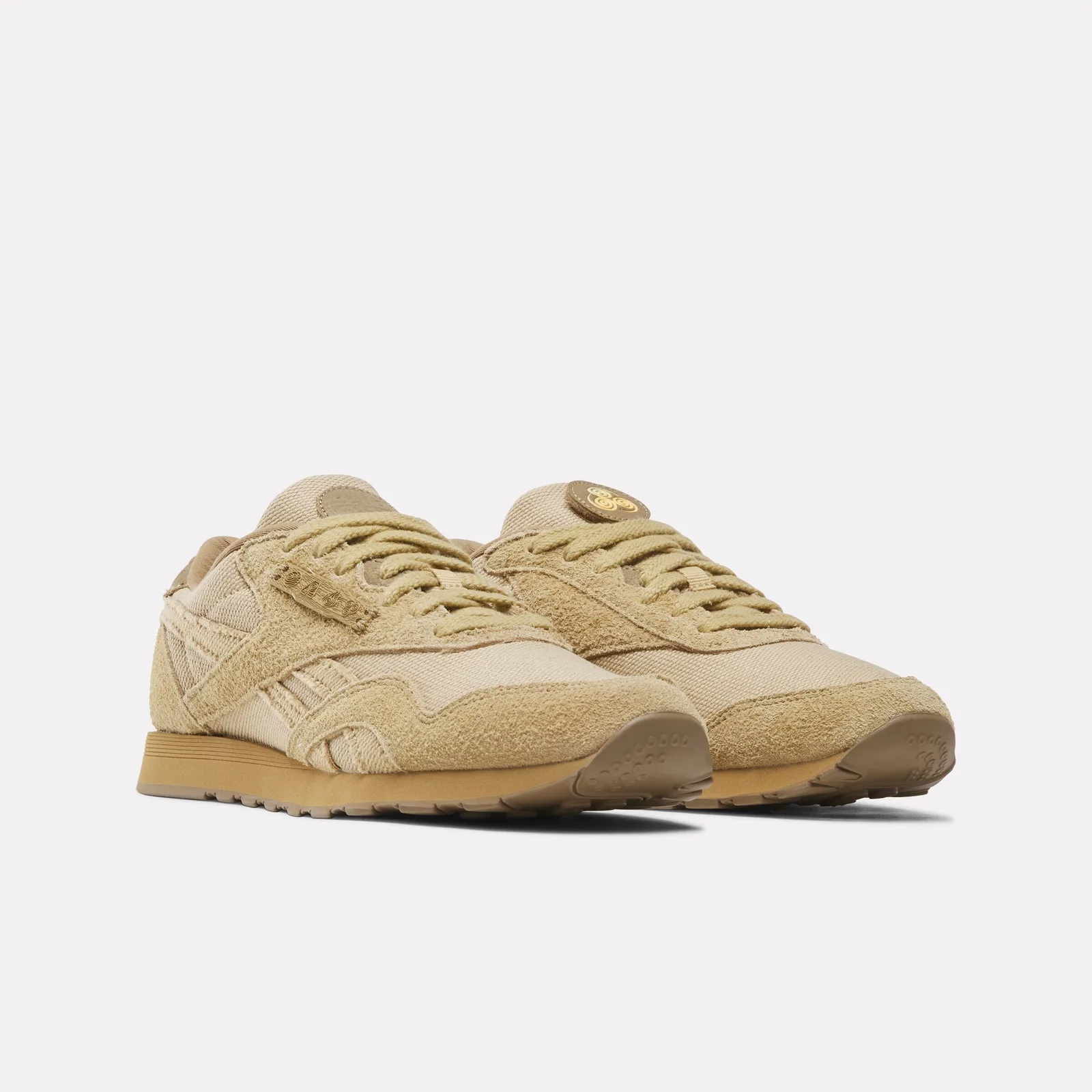 Front three-quarter pair in tan suede with a round gold swirl badge on one tongue, Four Nations