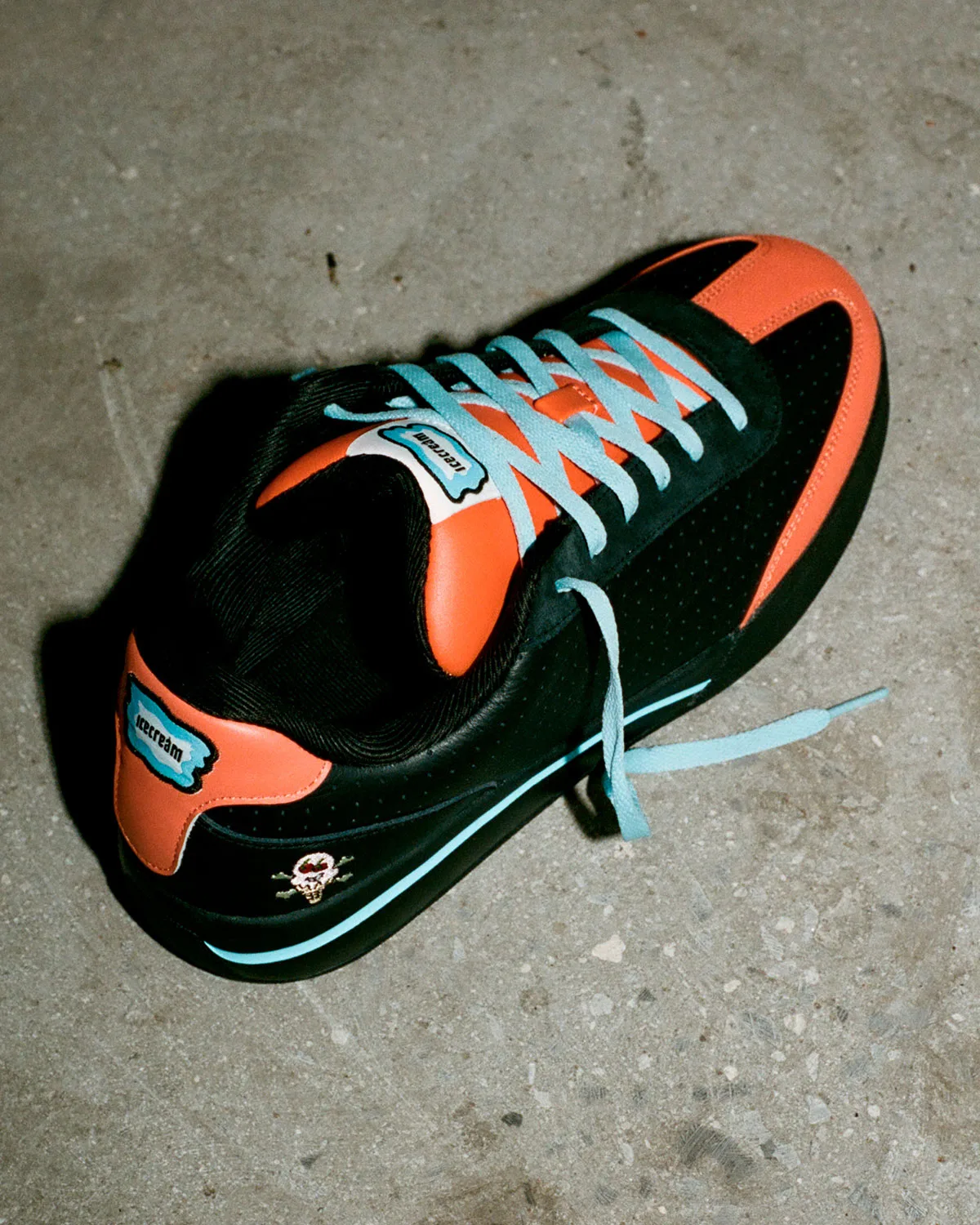 Top-down angled single shoe on concrete, black upper, orange heel tab, light blue laces, skull ice cream cone logo, Pumpkin