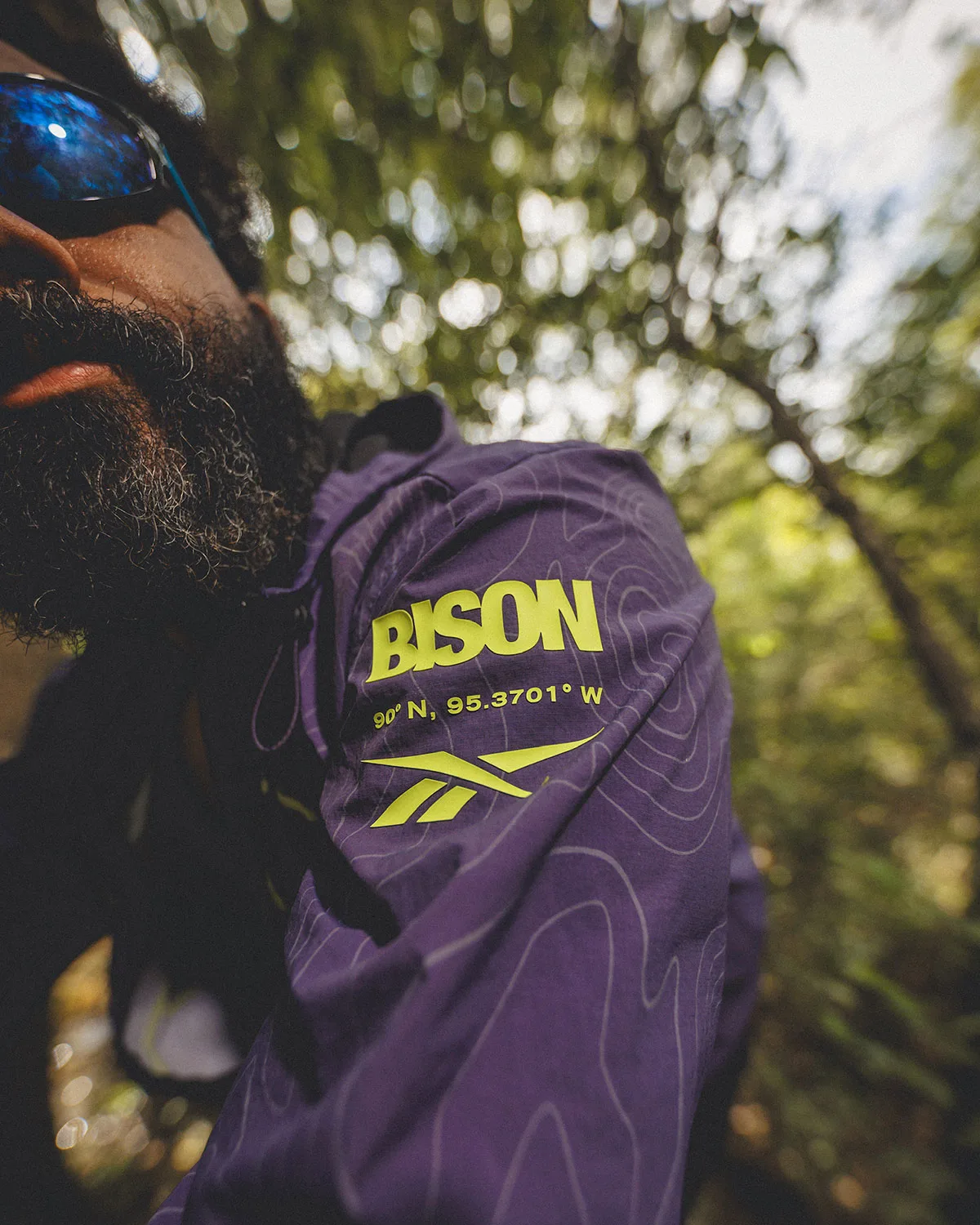 Man in sunglasses wearing purple topographic jacket with yellow BISON text and Reebok logo, forest, Reebok DMX Trailzilla