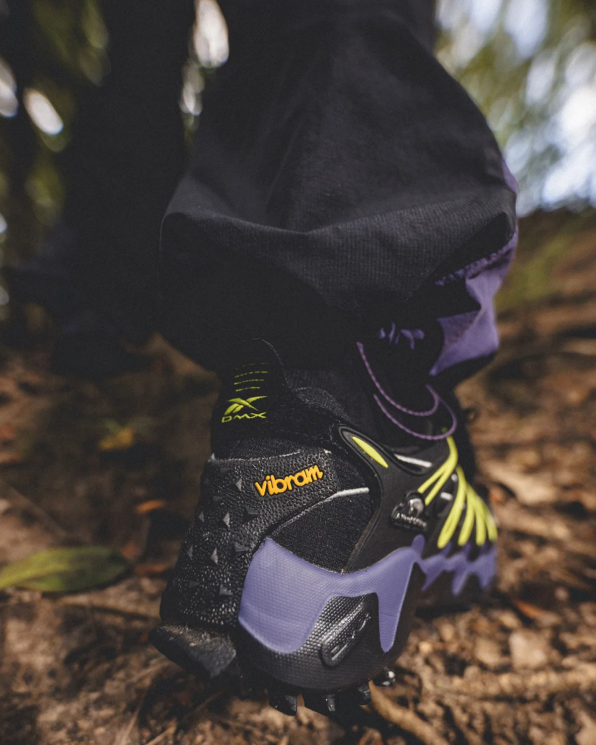 Rear on-foot view on dirt trail, black trousers, DMX heel logo, orange Vibram patch, Reebok DMX Trailzilla