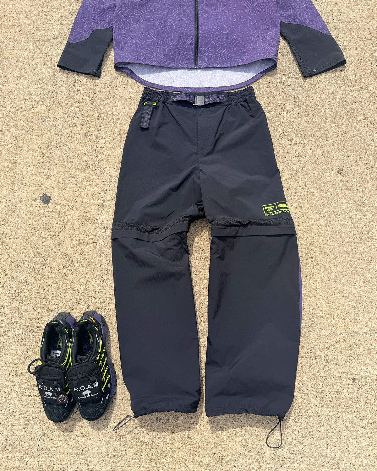 Flat lay of black zip-off pants and purple jacket, pair of shoes at lower left, Reebok DMX Trailzilla