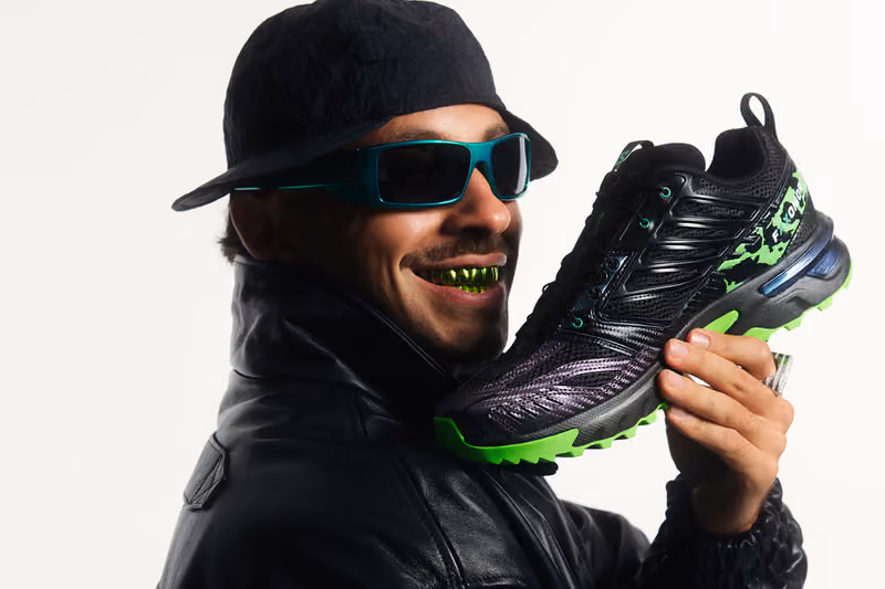 Feid holding the black and green ACS NEXXT beside his face while wearing sunglasses and a cap