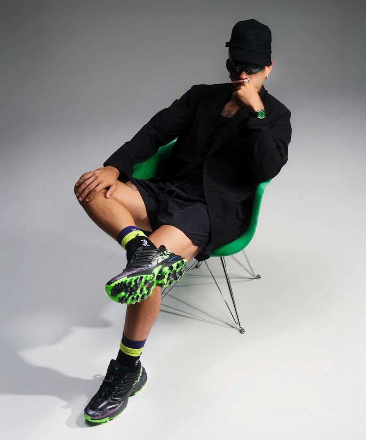 Model seated on a green chair wearing black clothing and the black-and-green Salomon pair