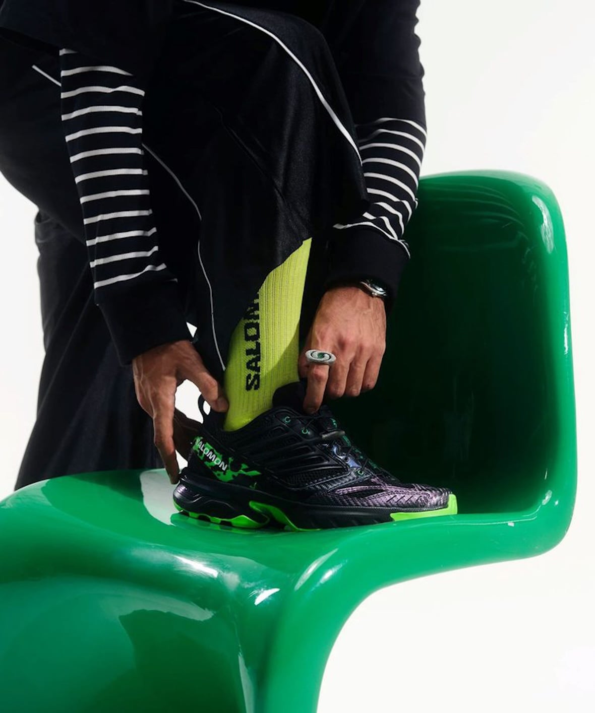 Close-up of a seated model's hands, striped sleeves and yellow-green Salomon sock beside a green chair